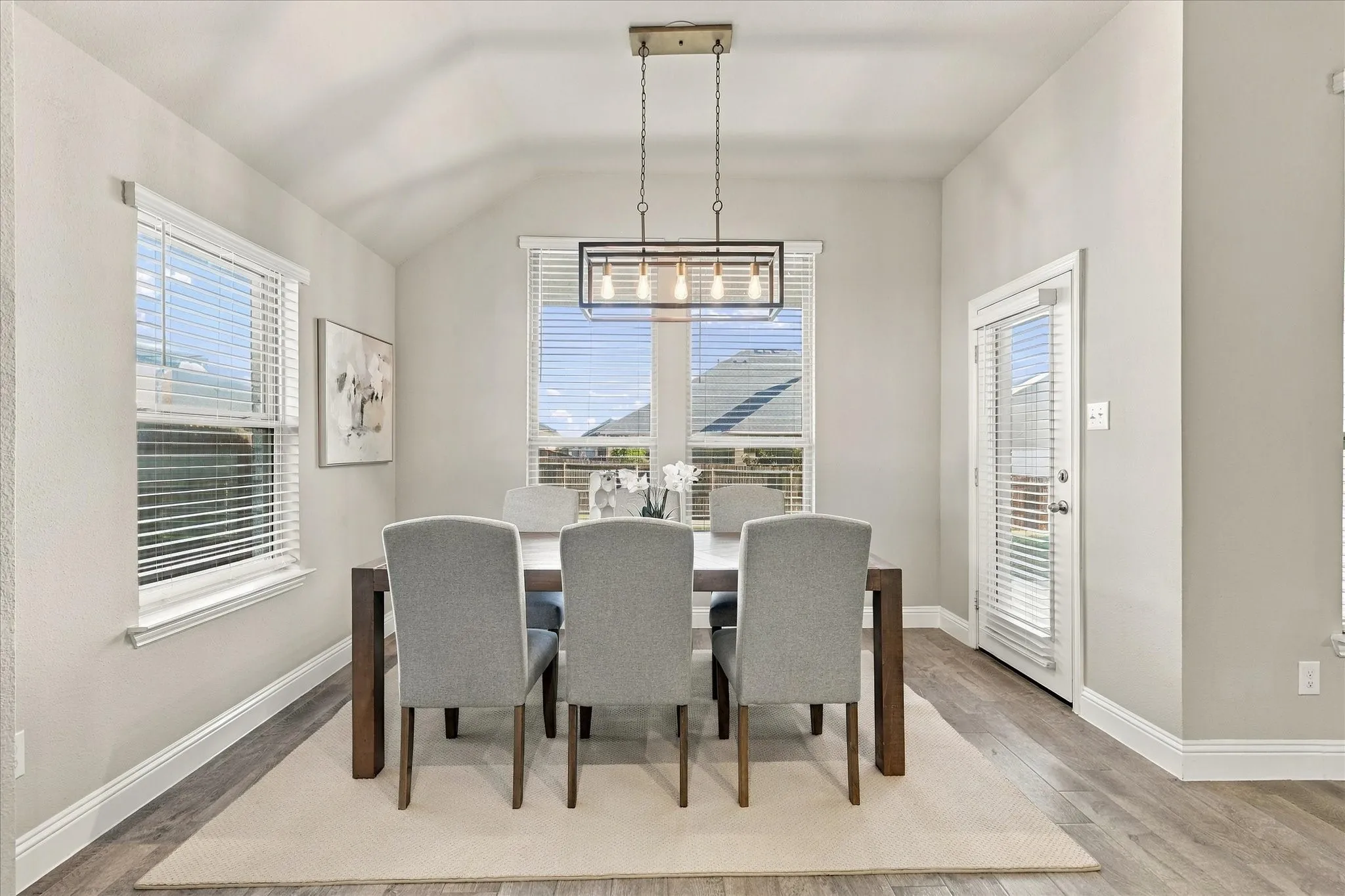 Vaulted ceiling, wood-finish flooring, two windows with blinds, a glass-paneled door, and a rectangular hanging light fixture