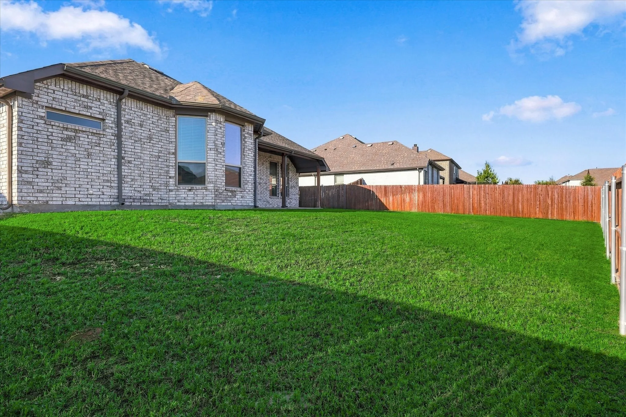 Expansive green lawn with a wood fence, brick exterior, multiple windows, and a shingle roof