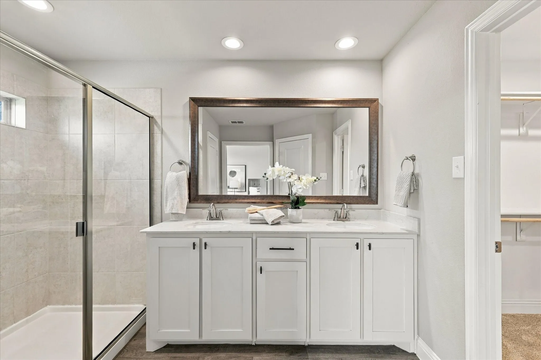 Dual vanity with white cabinetry and integrated sinks, large framed mirror, glass-enclosed shower with tiled walls, recessed lighting, and wood-finish flooring
