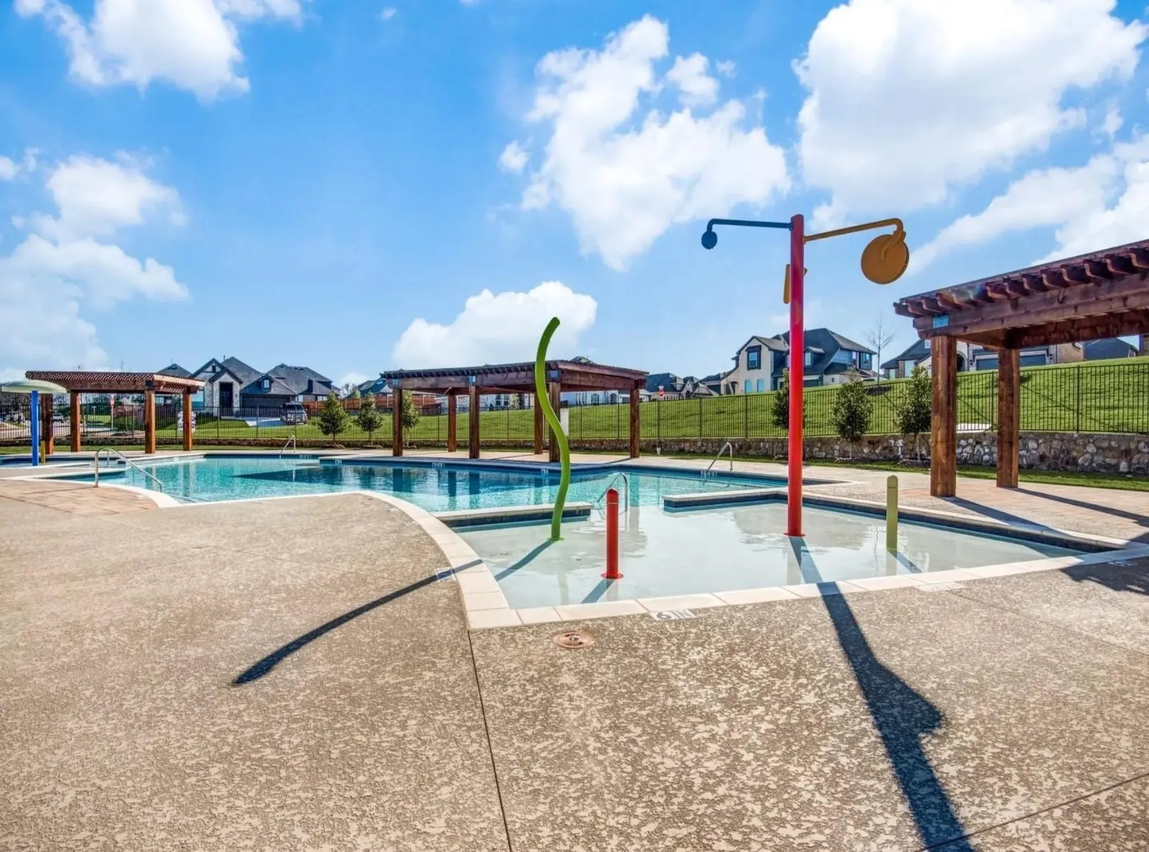 Large pool area featuring multiple wooden pergolas, a splash pad with water features, and concrete decking