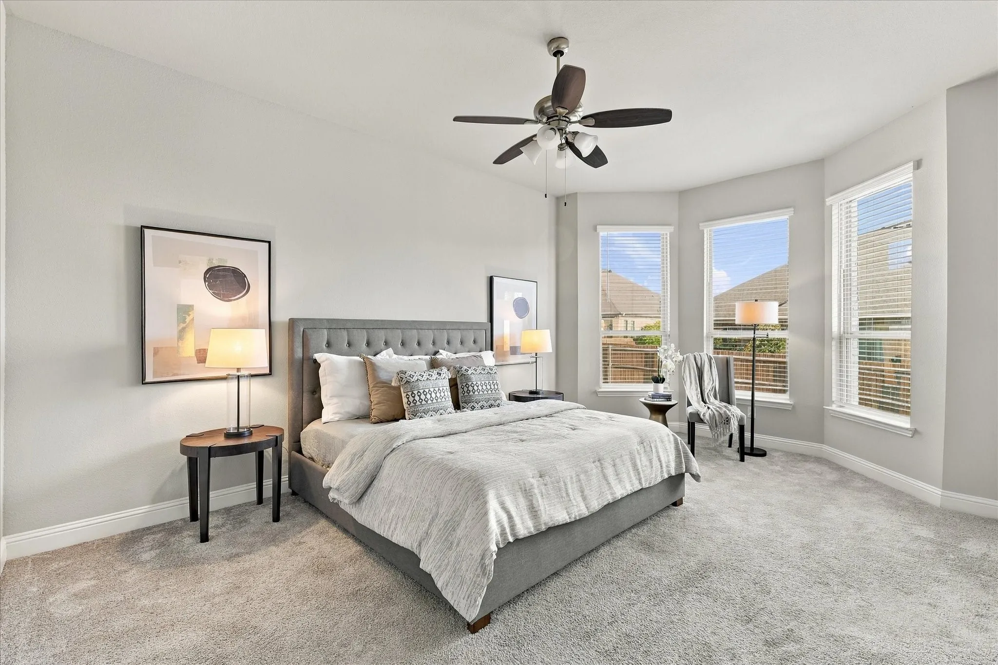 Bay window feature with three large windows, light gray walls, light gray carpet flooring, ceiling fan with integrated lighting, and a tufted upholstered bed frame
