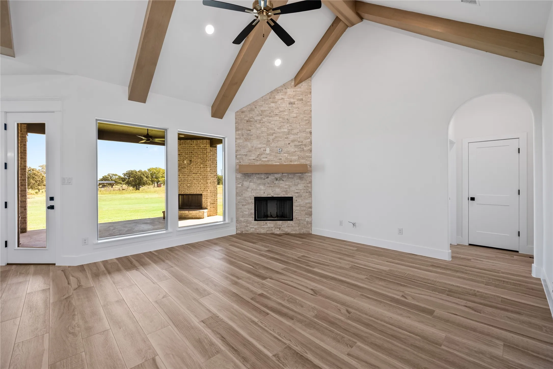 Vaulted ceiling with exposed wood beams, stone-faced fireplace with wood mantel, large fixed-pane windows, wood-finish flooring, and an arched entryway