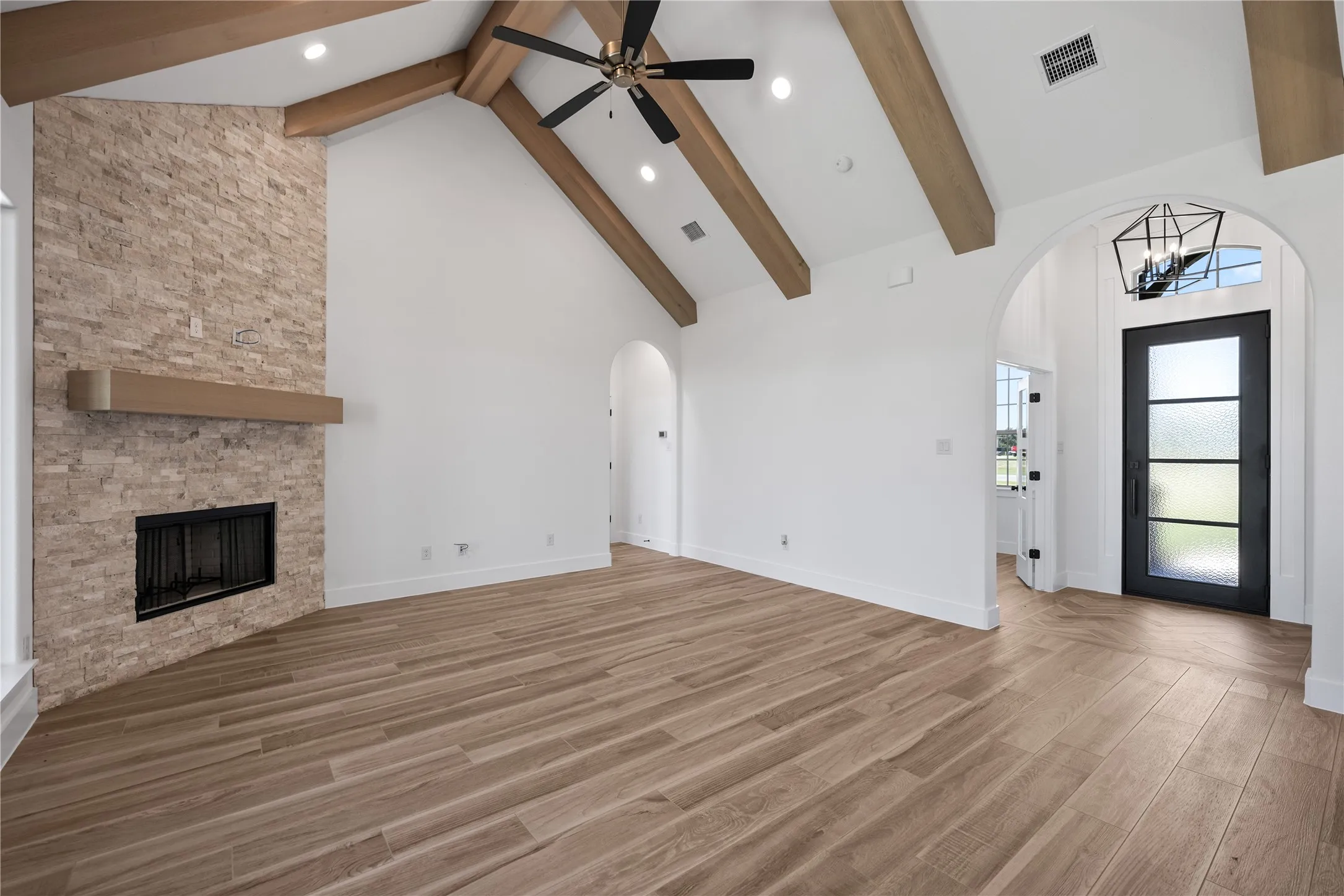 Vaulted ceiling with exposed wood beams, stone-clad fireplace with mantel, wood-finish flooring, recessed lighting, and an arched entry with a glass-paneled door