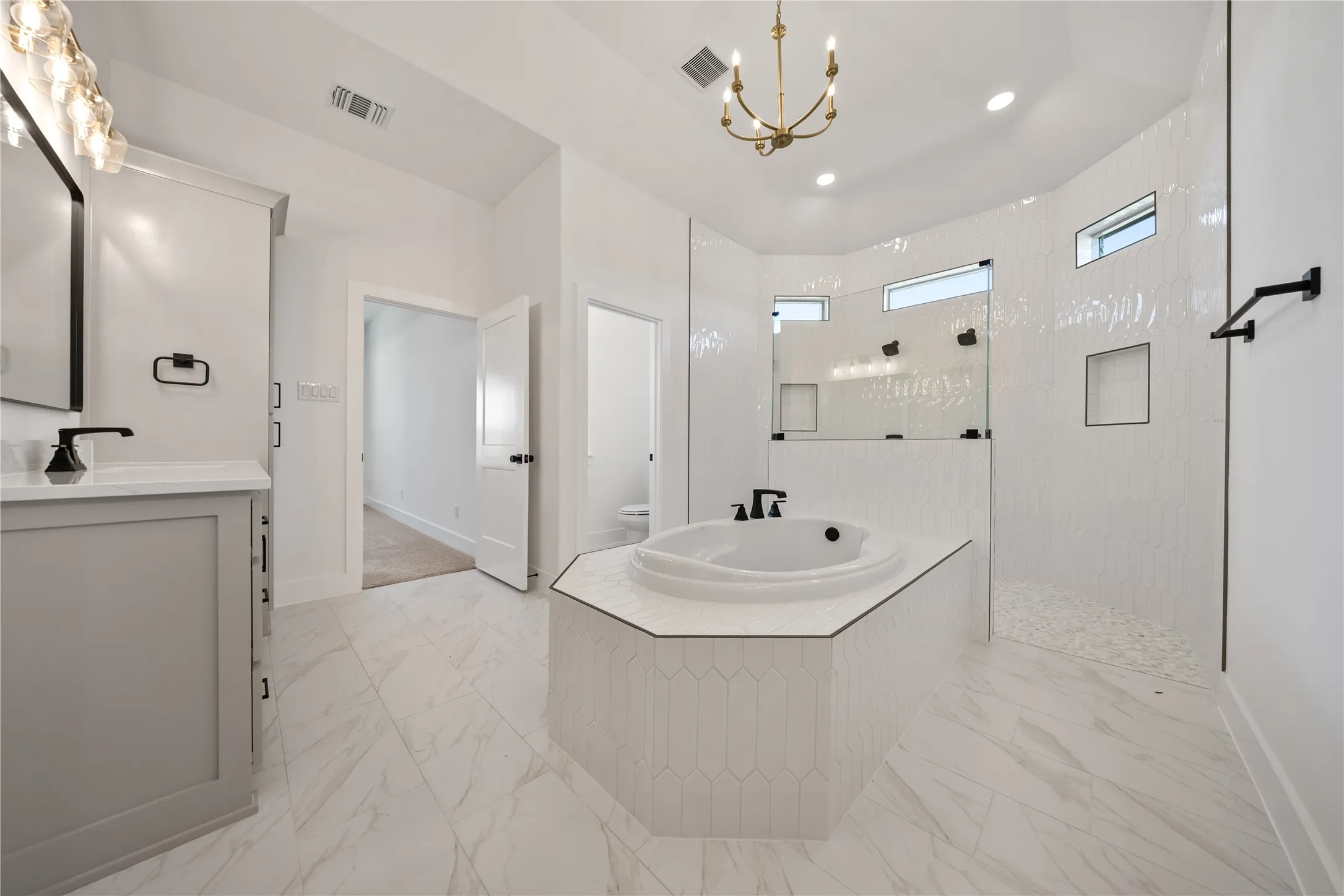 White tile flooring, built-in soaking tub, walk-in shower with glass enclosure, single vanity with white countertop, and brass chandelier