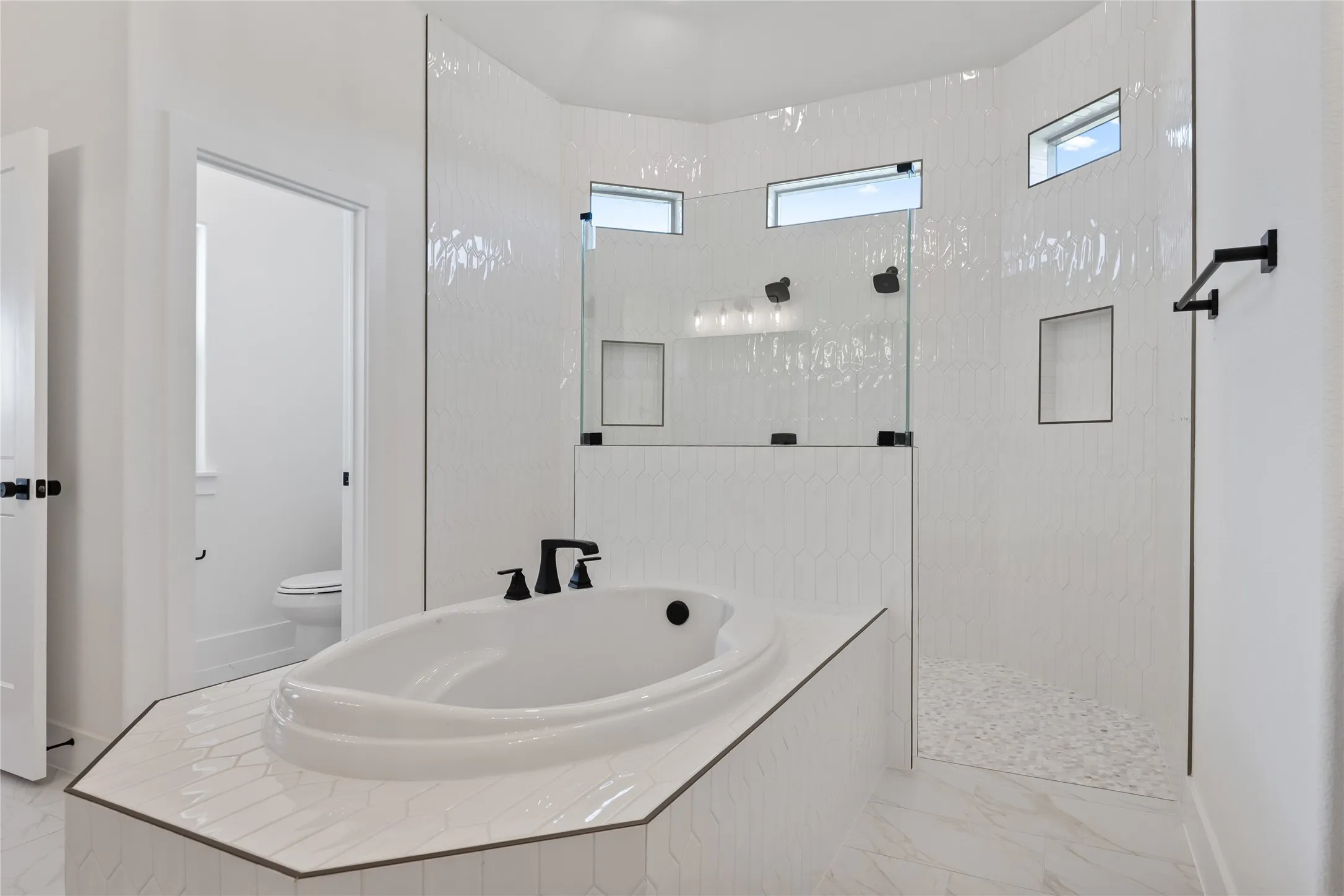 Soaking tub with matte black fixtures, an expansive walk-in shower with glass enclosure, white hexagon tile walls, and large format floor tile