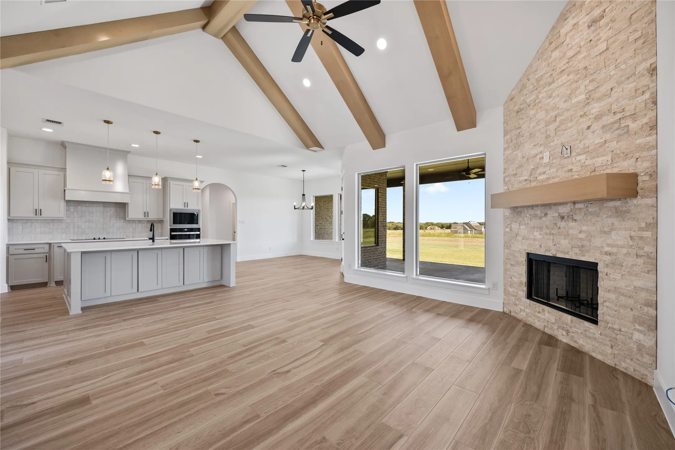 Vaulted ceiling with exposed wood beams, wood-finish flooring, stone-clad fireplace with wood mantel, large windows, and a kitchen island with pendant lighting