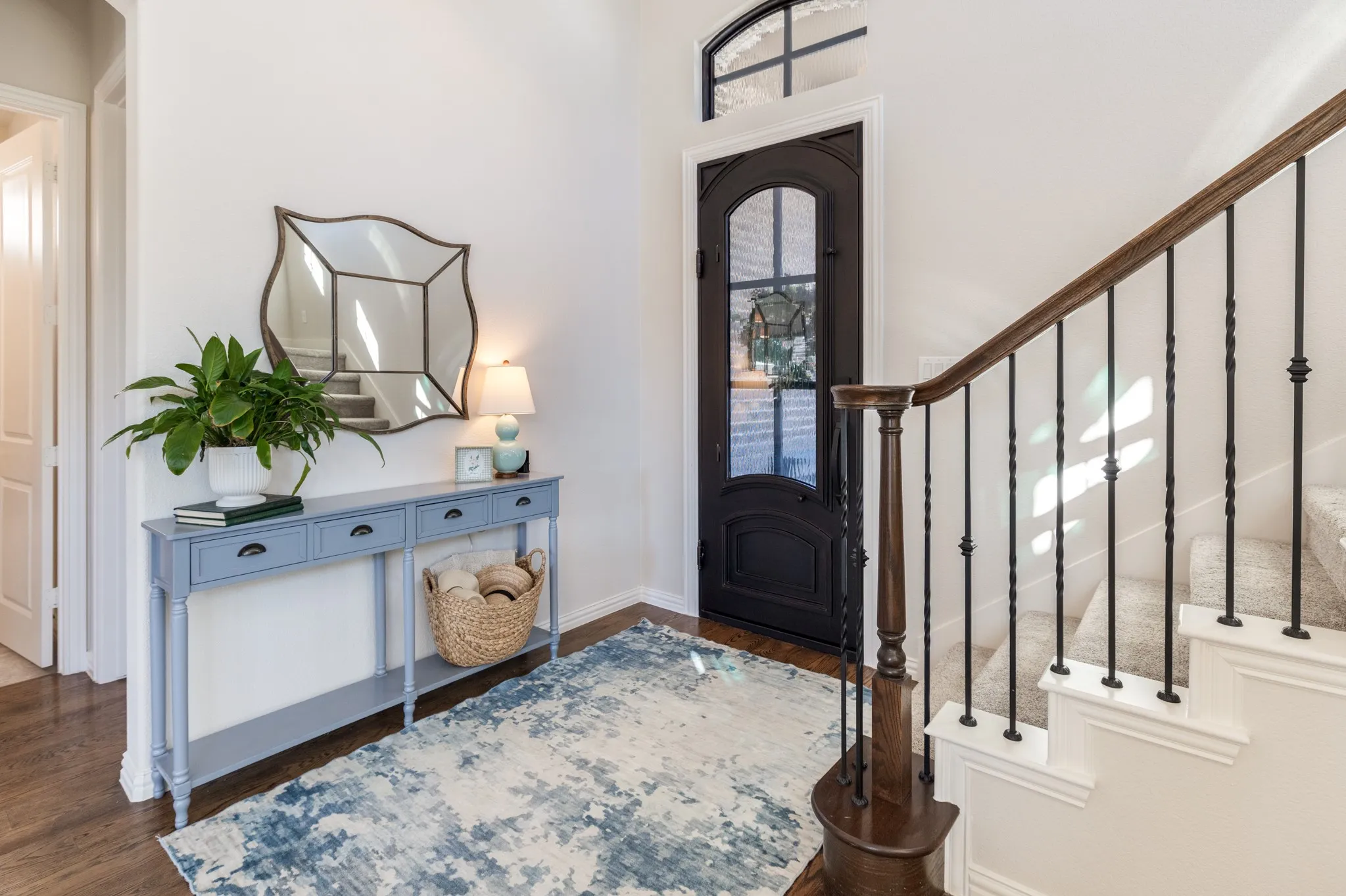 Arched entryway door with glass paneling, overhead transom window, wood-finish flooring, and a carpeted staircase with wrought iron balusters and a wood handrail