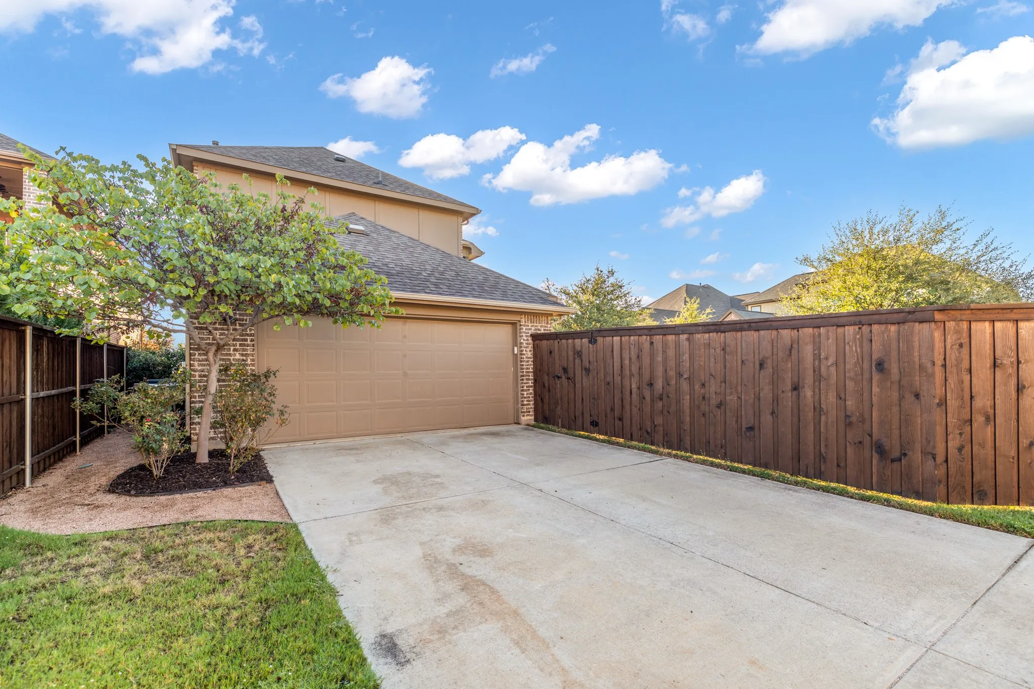 Attached garage with a tan door, brick facade, concrete driveway, and wood privacy fencing