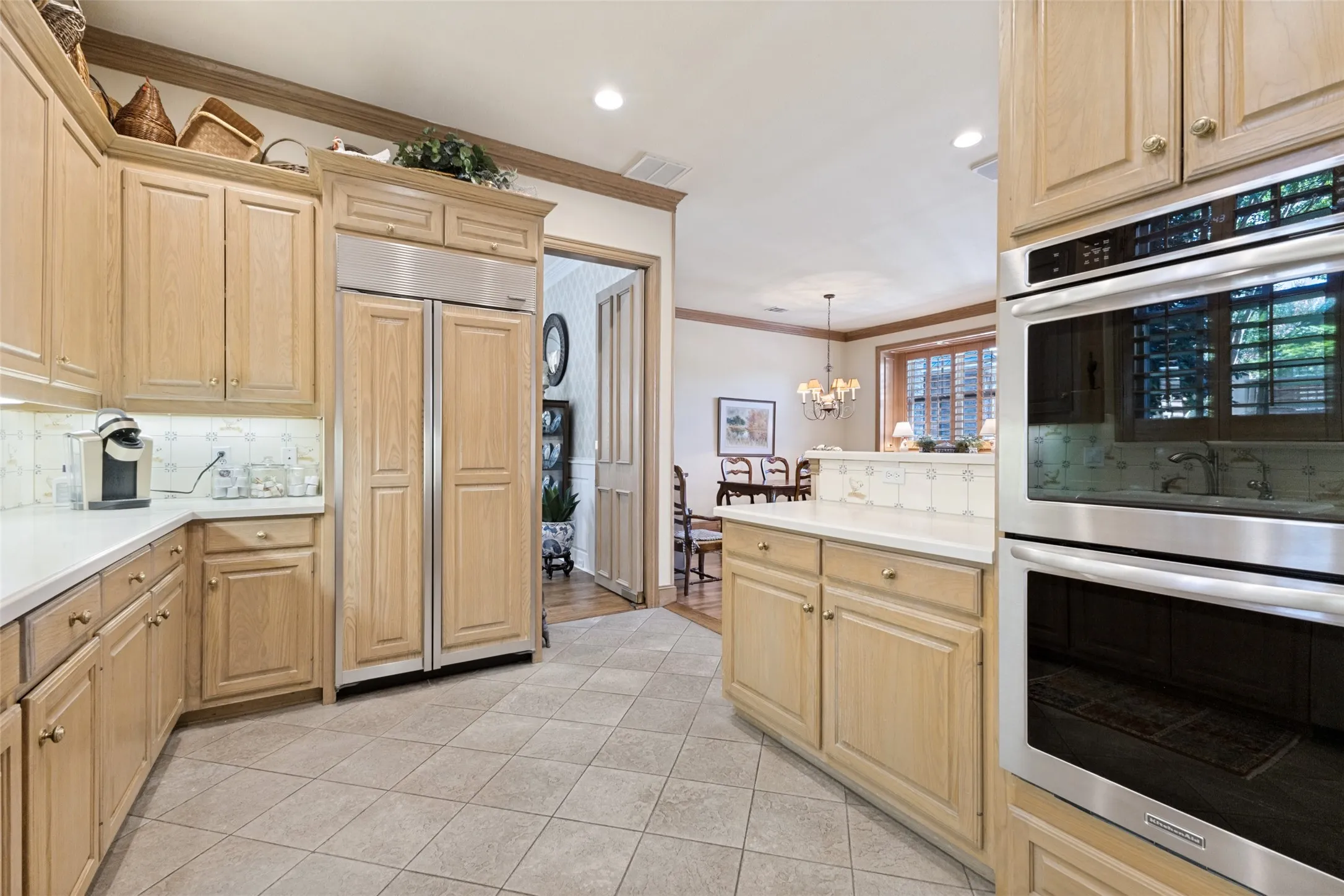 Light wood-finish cabinetry with gold-tone hardware, integrated stainless steel refrigerator, double wall ovens, white countertops, and tiled flooring