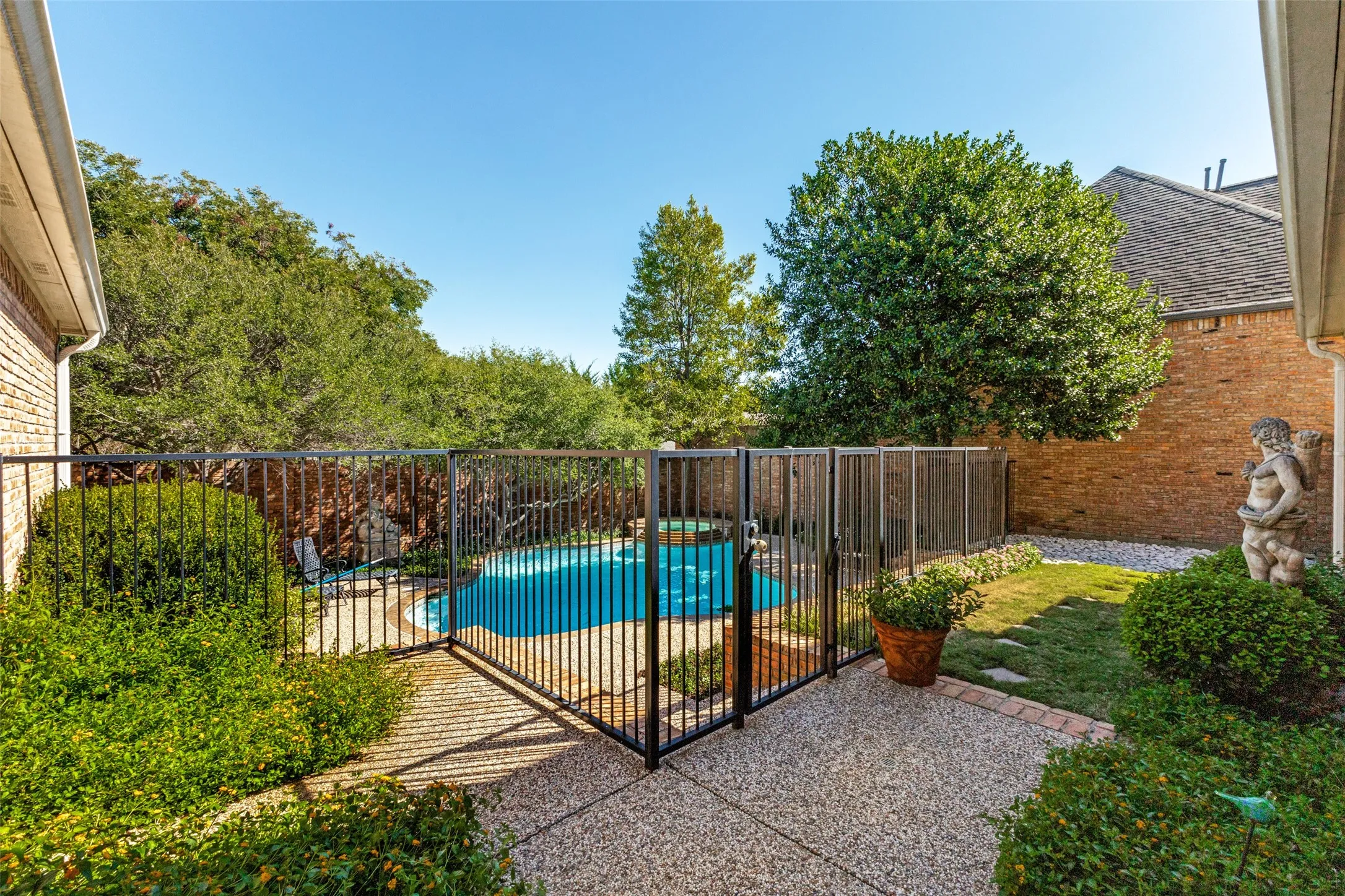 Pebbled concrete patio with a dark metal fence enclosing a pool, surrounded by mature landscaping and a brick-clad exterior