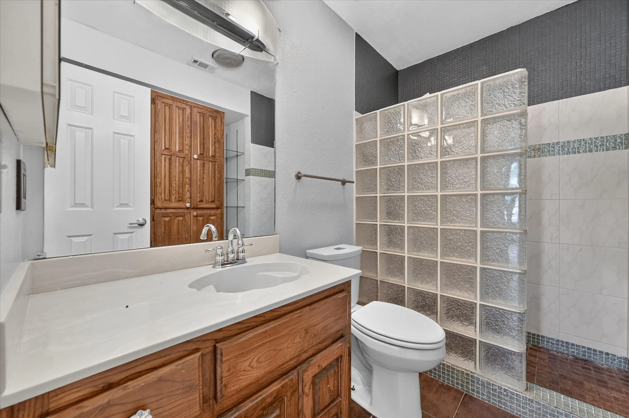 Vanity with an integrated sink and wood cabinetry, expansive mirror, glass block partition, walk-in shower with tile surround, and tile flooring