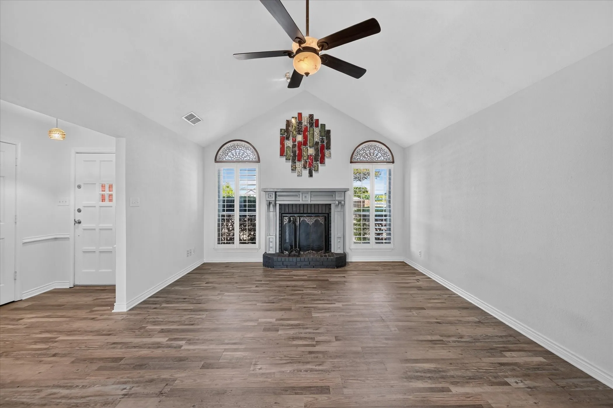 Vaulted ceiling with a ceiling fan, fireplace with a stone hearth, arched windows with plantation shutters, and wood-finish flooring