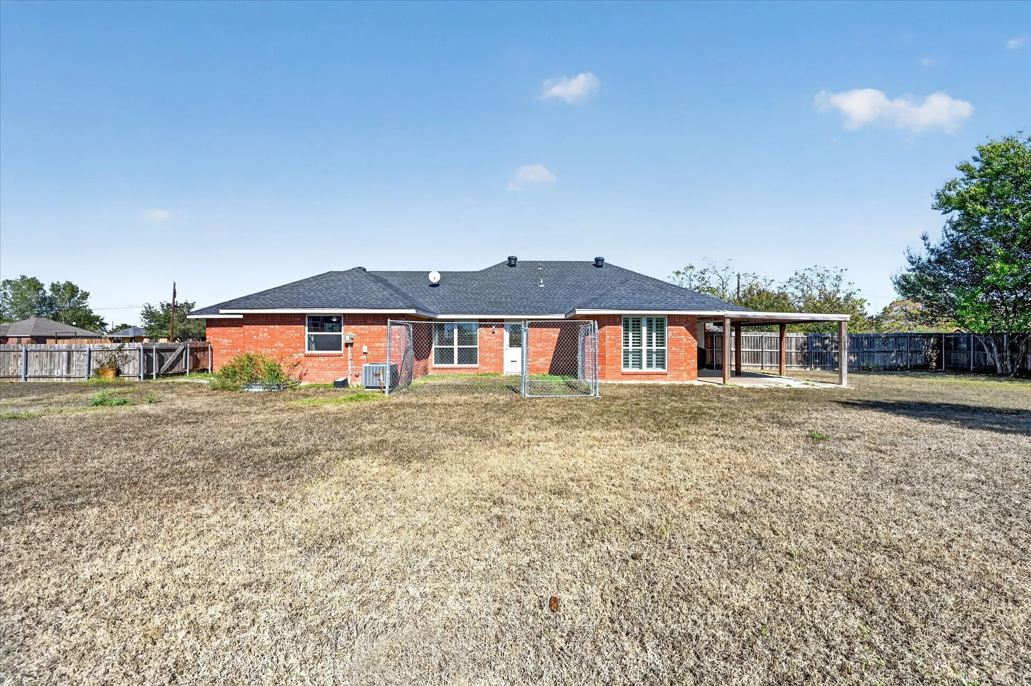 Red brick exterior, dark shingle roof, covered patio structure, fenced yard perimeter