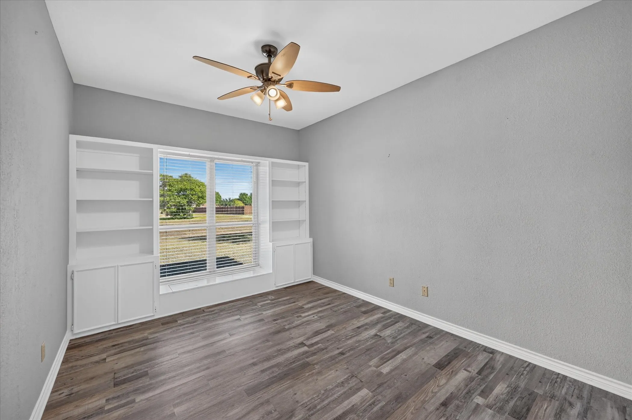 Wood-finish flooring, built-in shelving, window with blinds, ceiling fan with light fixture