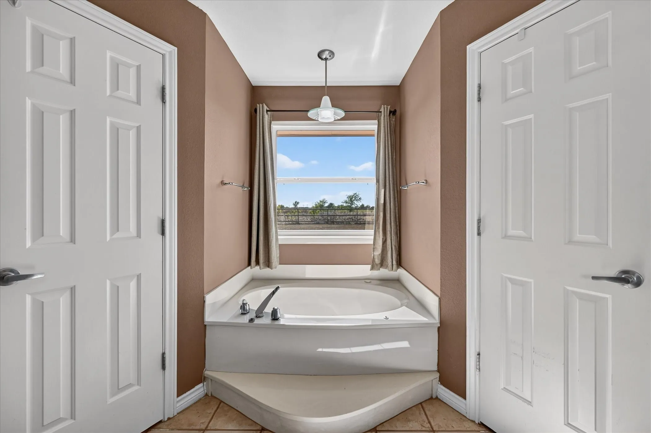 Built-in soaking tub with a surrounding white countertop, under a window with drapes, complemented by a suspended light fixture