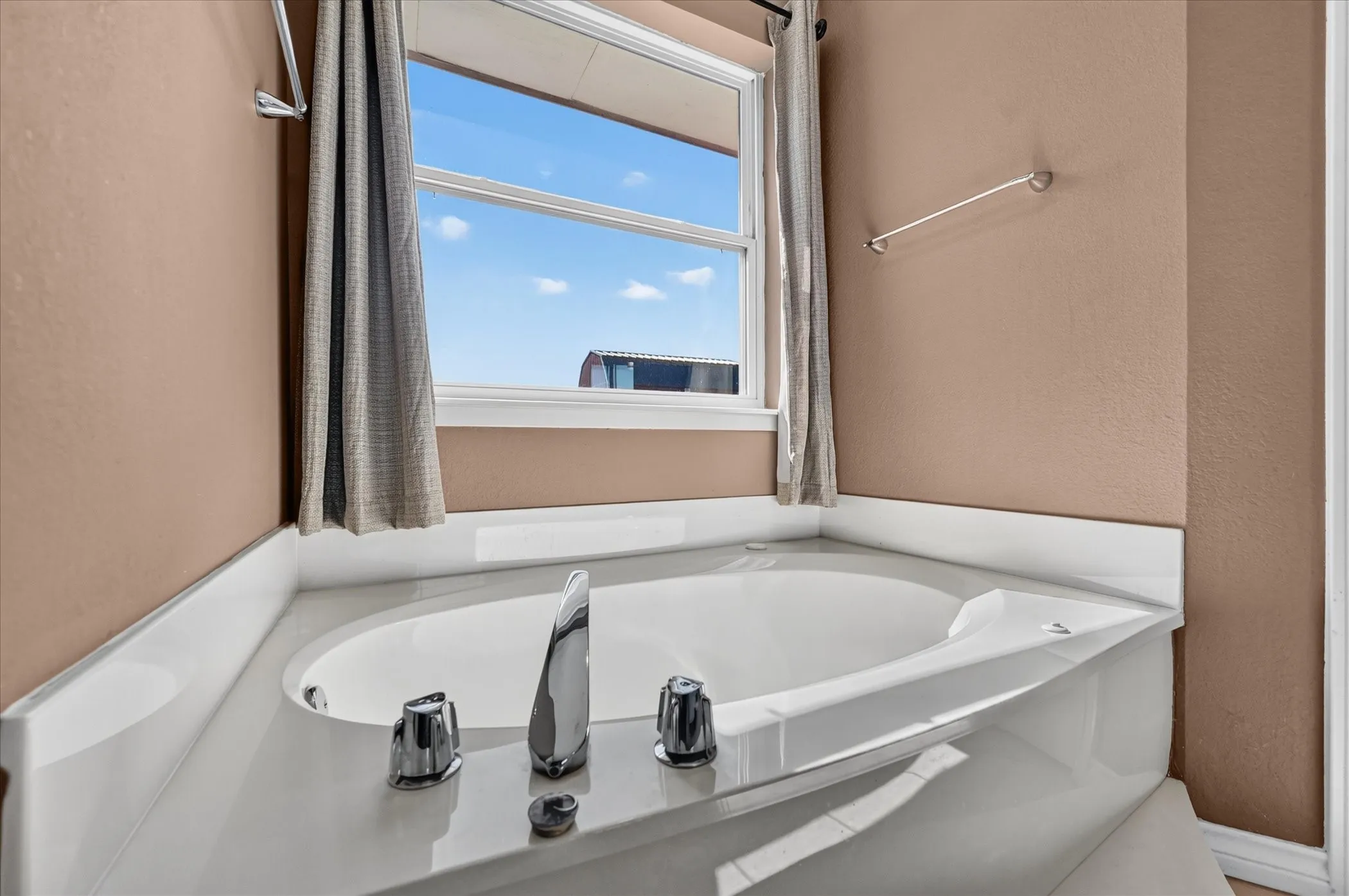 Corner soaking tub with deck-mounted chrome fixtures, set against a window with a white frame and sill