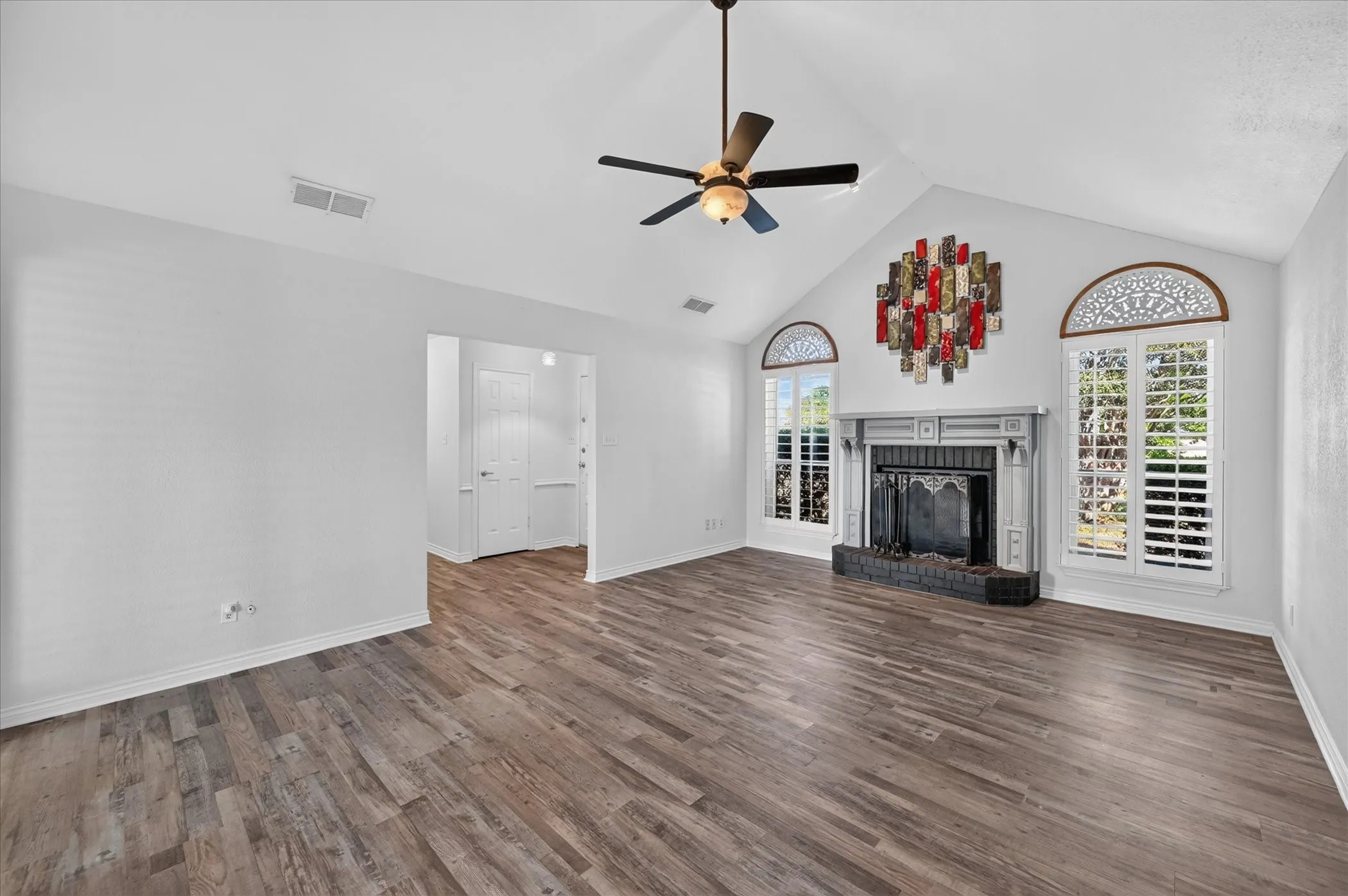 Vaulted ceiling with a ceiling fan, fireplace with a mantel, wood-finish flooring, and arched windows with plantation shutters