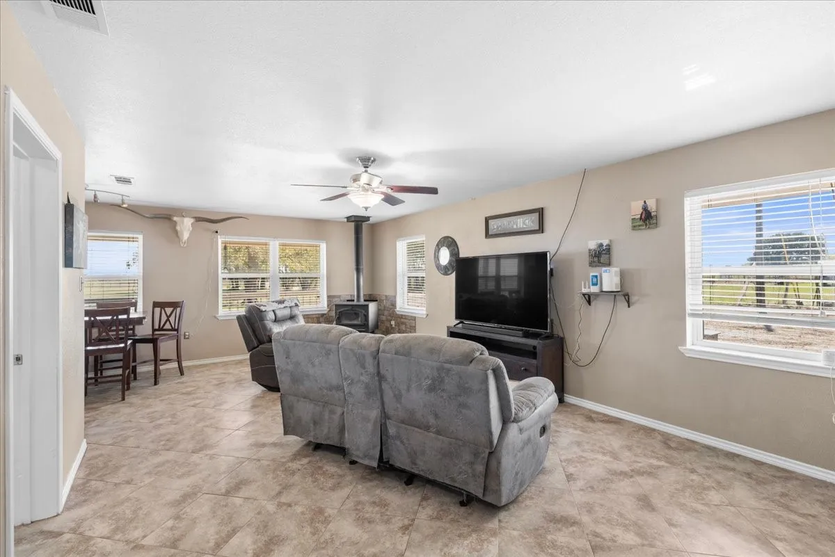 Open-concept interior featuring a wood-burning stove with stone-finish surround, multiple windows with blinds, a ceiling fan, and tile flooring