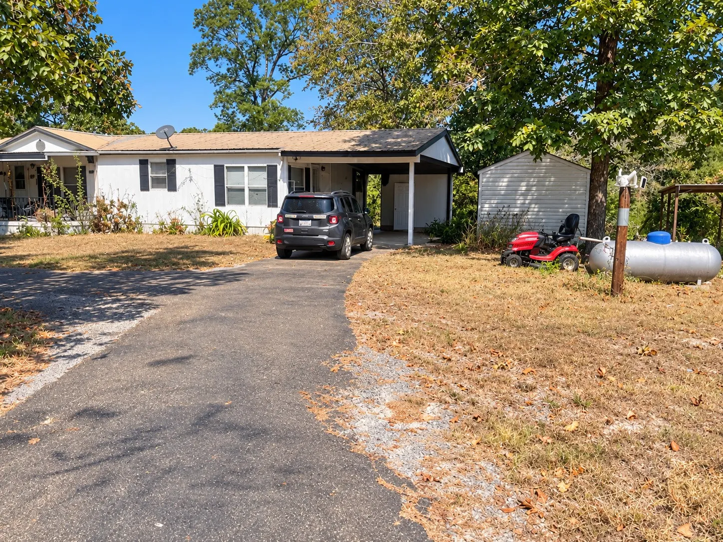 White siding exterior with dark shutters, covered porch entry, covered carport, detached storage shed, and a paved driveway