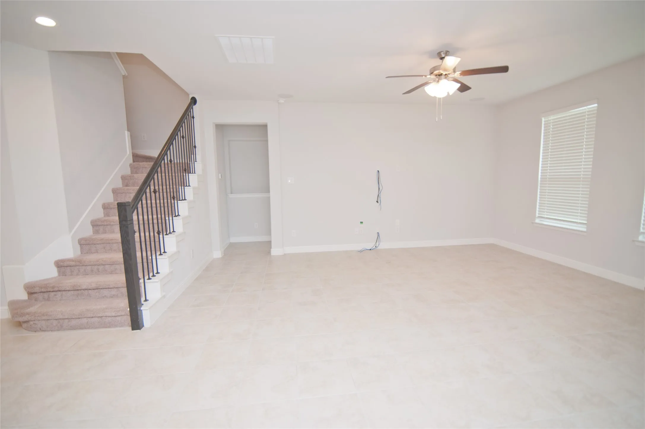 Spacious room featuring tile flooring, a staircase with carpeted treads and wrought iron balusters, a ceiling fan with integrated lighting, and multiple windows with blinds