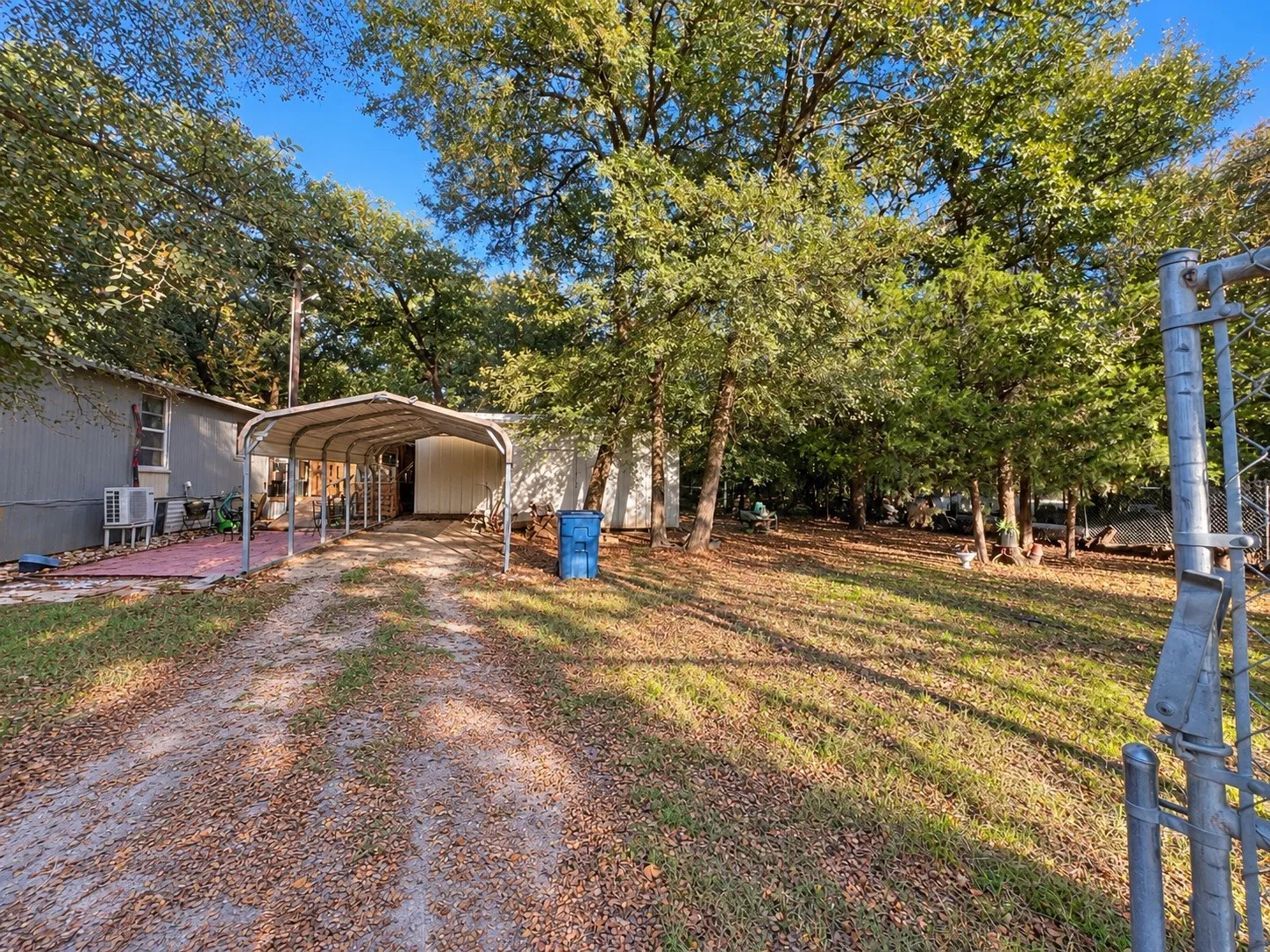 Gravel driveway leading to a metal carport structure, a side patio with brick pavers, and a spacious grassy area with mature trees