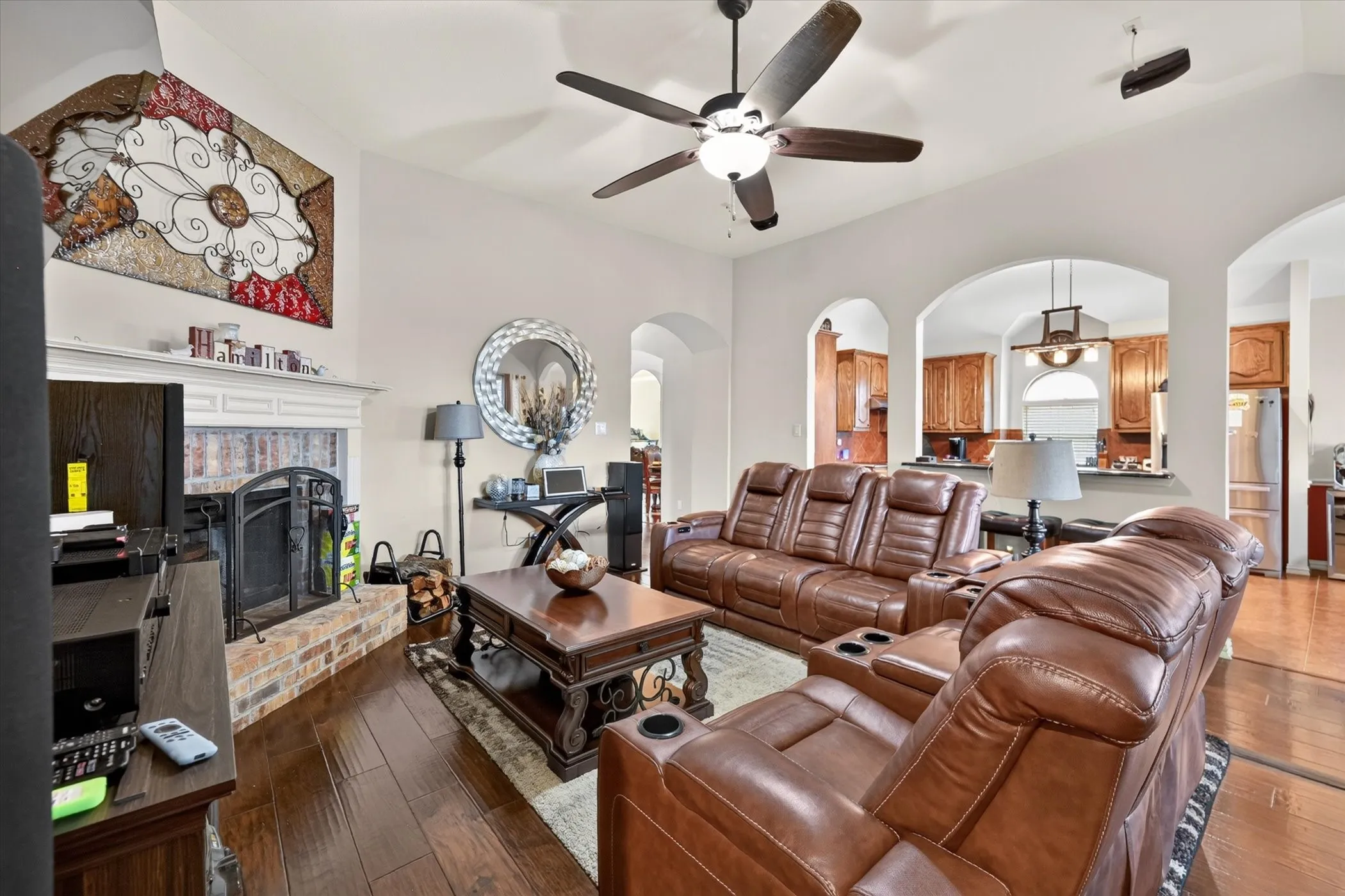 Spacious living area featuring wood-finish flooring, a ceiling fan, and a brick-surround fireplace with a white mantel