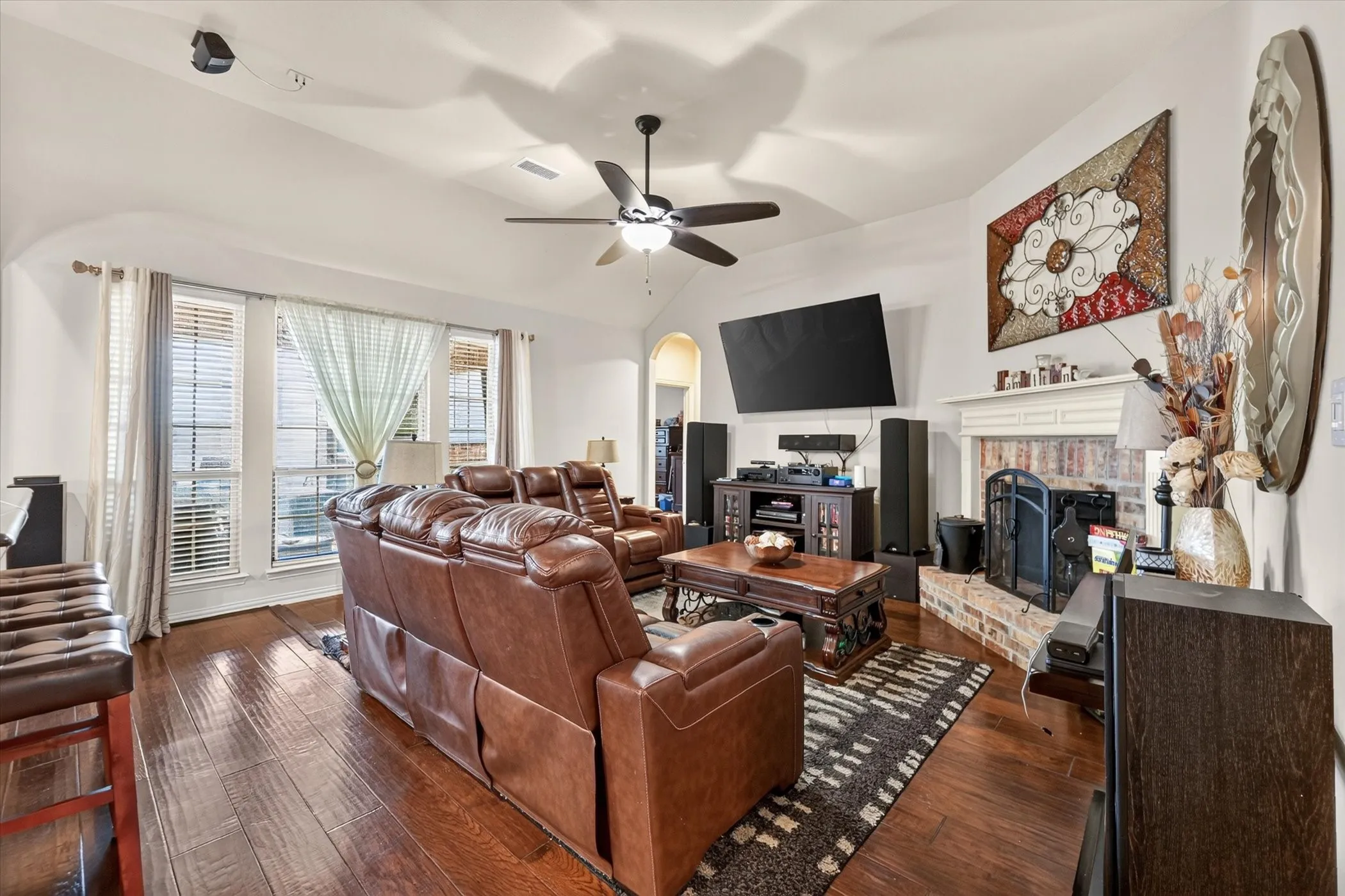 Vaulted ceiling with a ceiling fan, wood-finish flooring, multiple windows, and a brick fireplace with a white mantel