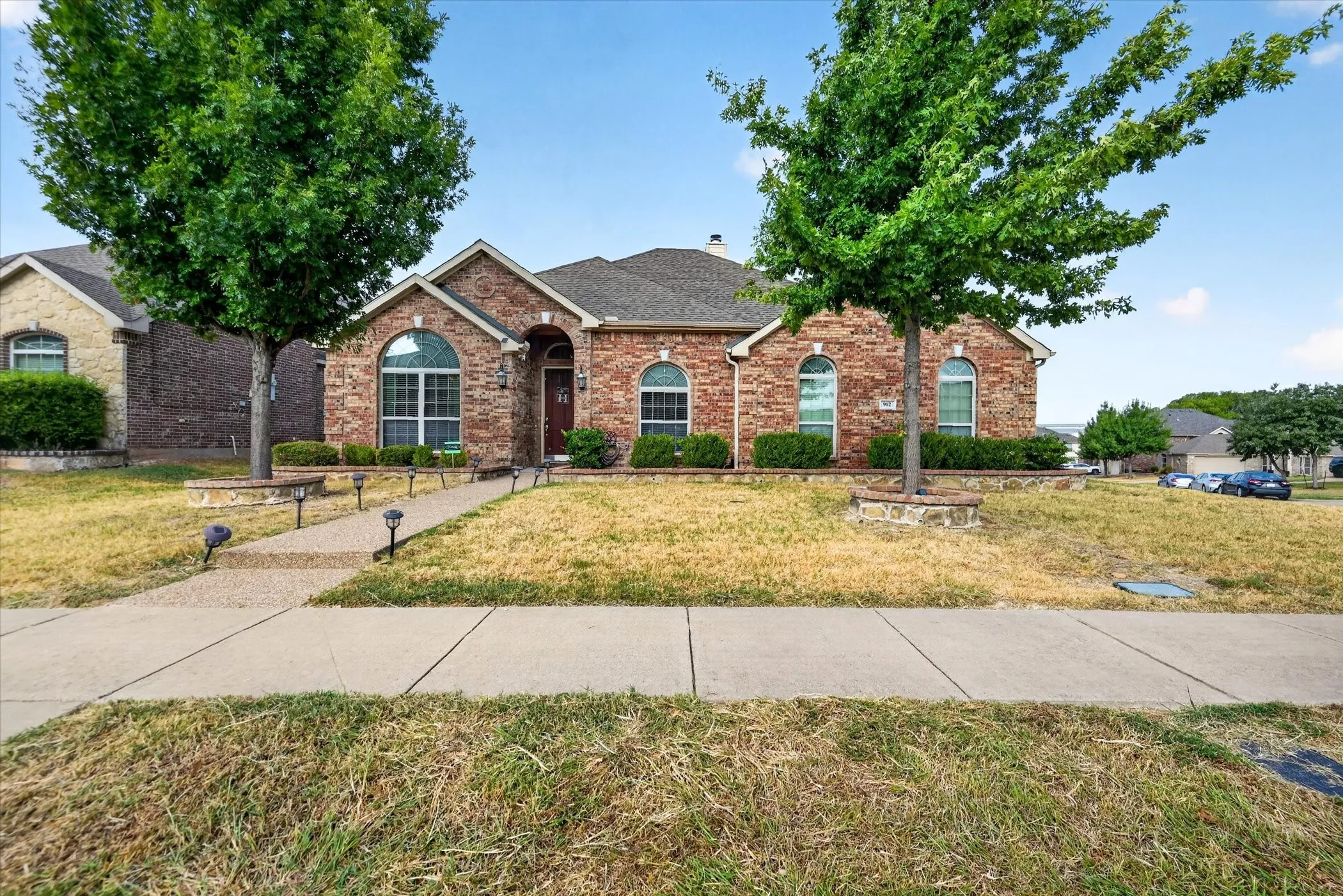 Brick facade with arched windows, mature trees, and a front walk with landscape lighting