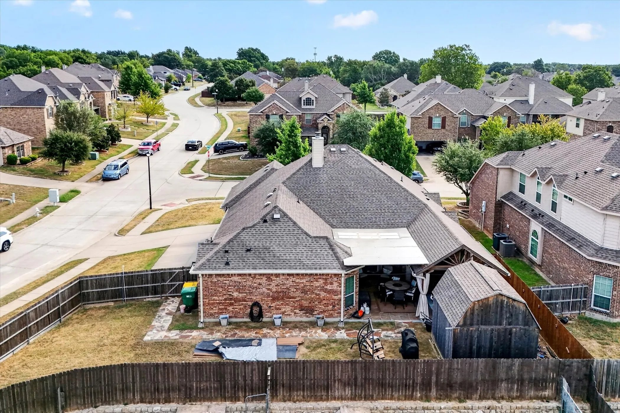 Red brick exterior with a dark shingle roof, featuring a covered patio, a detached shed, and a fenced backyard