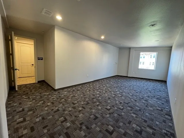 Spacious room with a patterned carpet, recessed lighting, and a white interior door with dark hardware