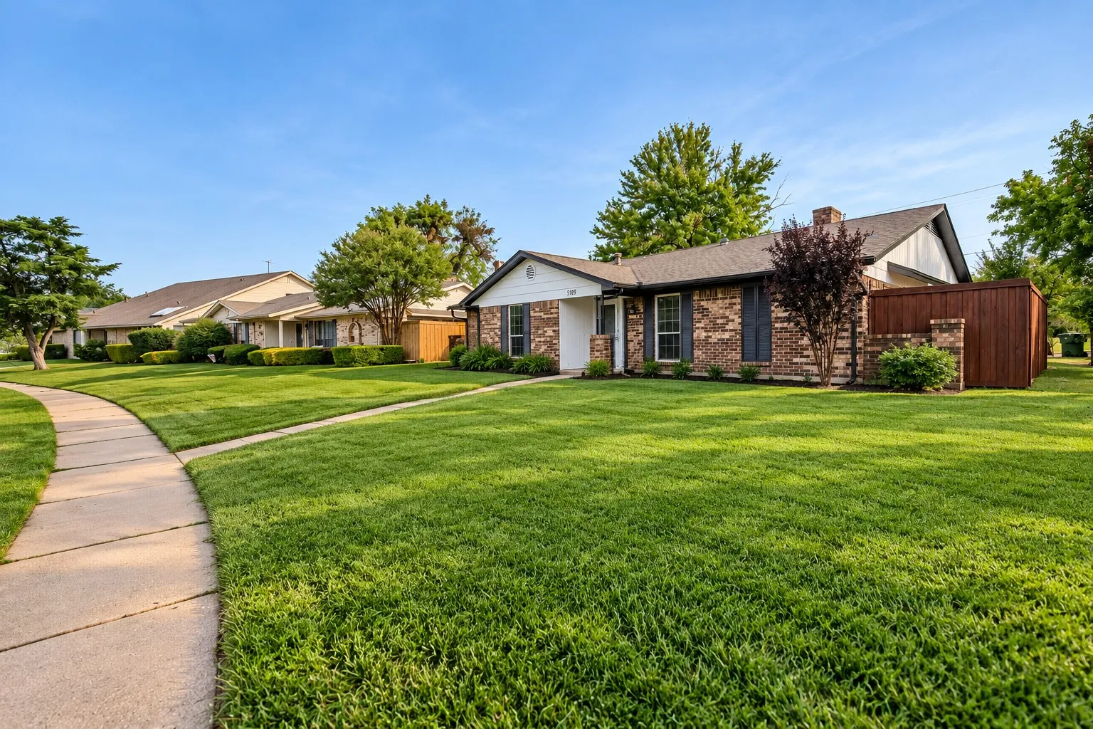 Brick facade home with a shingle roof, mature landscaping, and an adjacent privacy fence