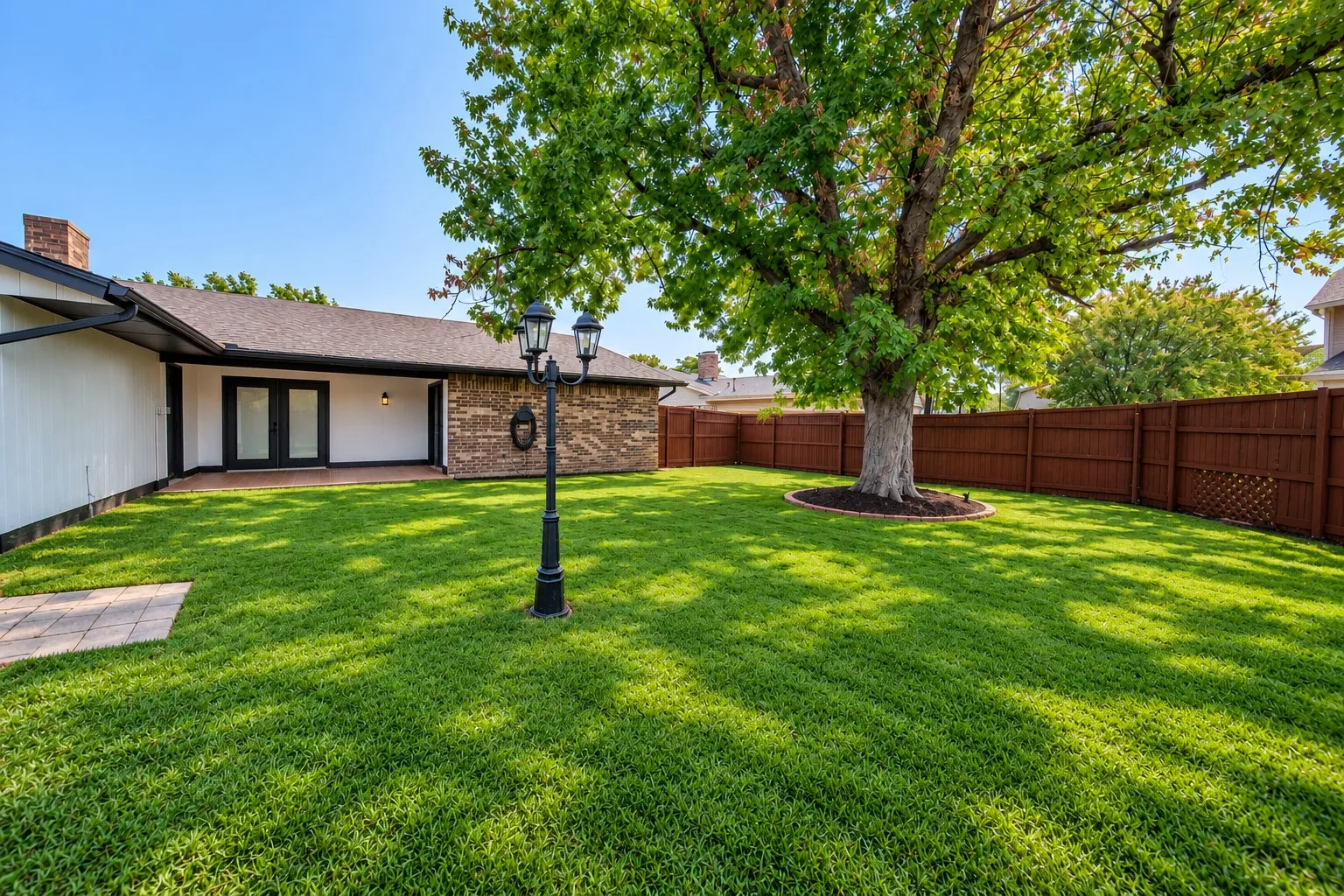 Expansive backyard featuring a large mature tree, wood privacy fencing, a brick exterior facade, and a covered patio with double doors