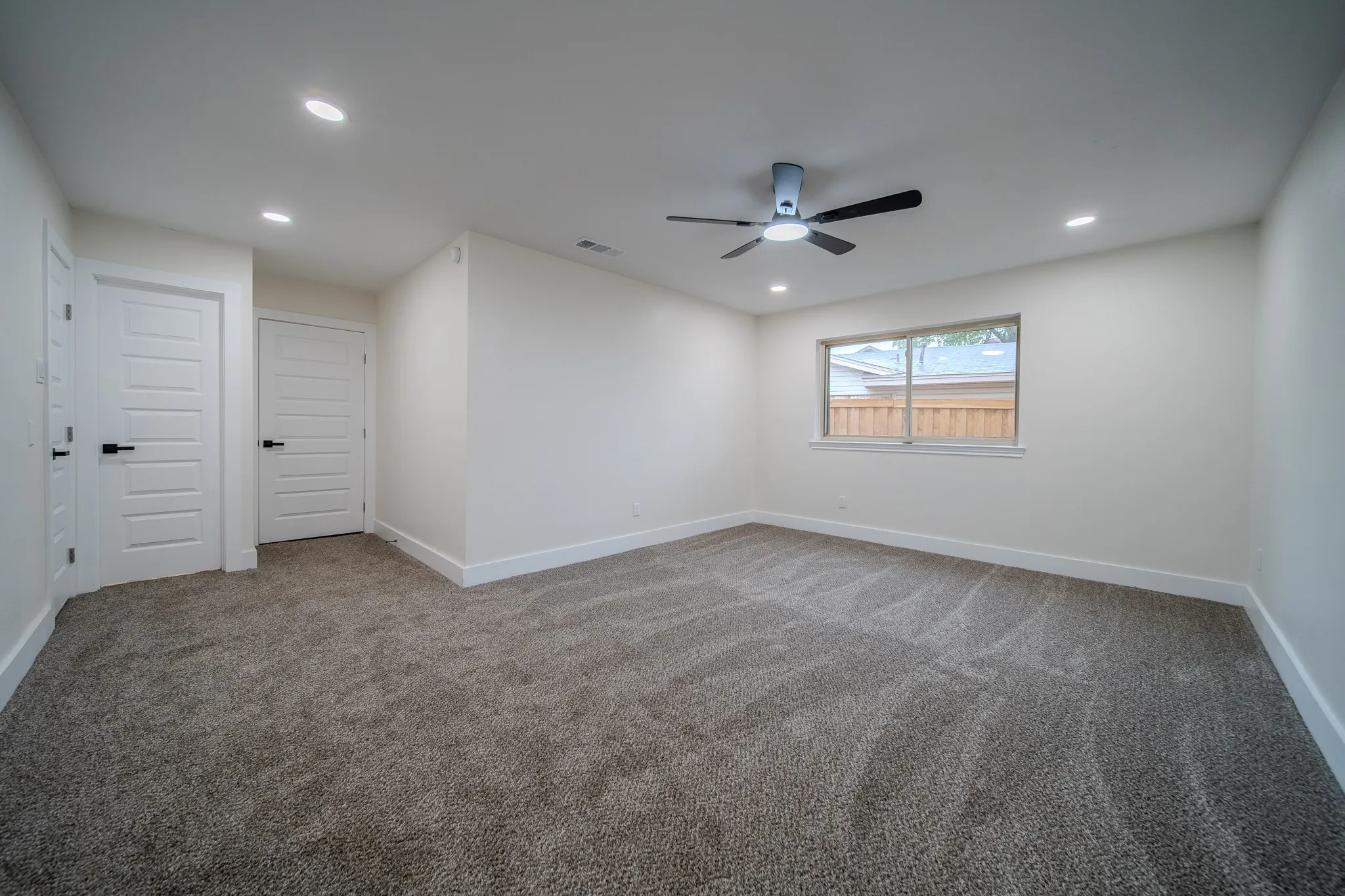 Spacious room with plush gray carpeting, recessed lighting, white panel doors with black hardware, a ceiling fan, and an exterior window
