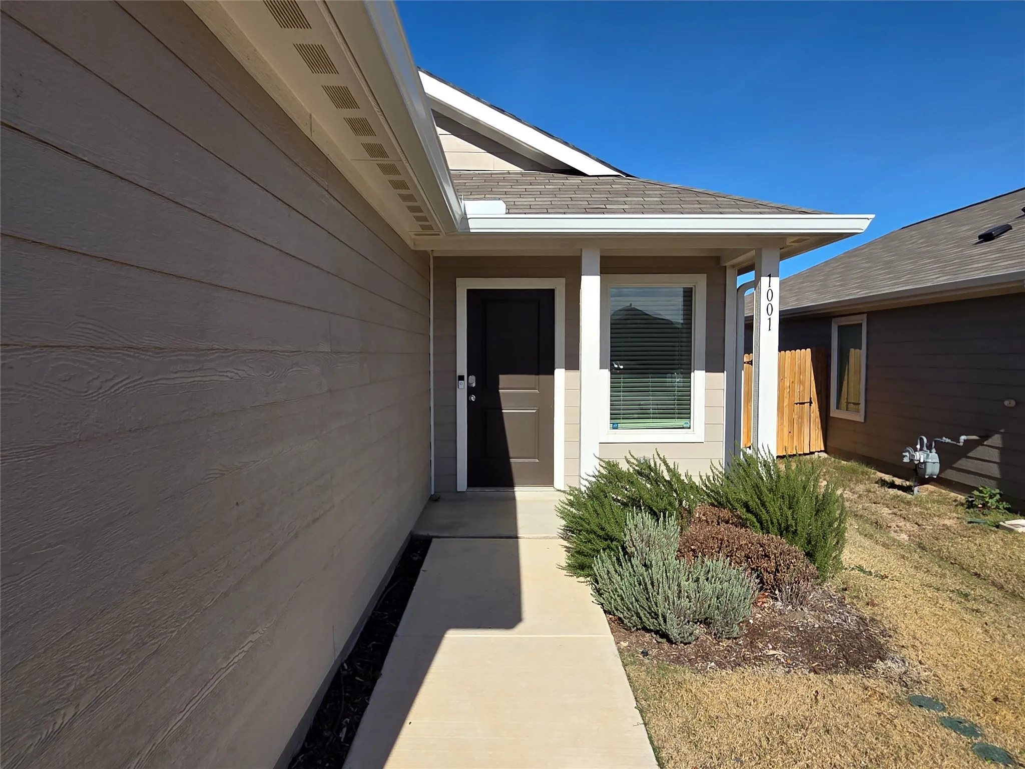 Tan siding with white trim, covered entryway, dark brown entry door, concrete walkway, and decorative landscaping