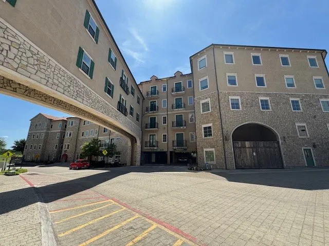 Multi-story facade featuring stucco and stone veneer, multiple balconies, a covered pedestrian bridge, arched entryway, and paver driveway