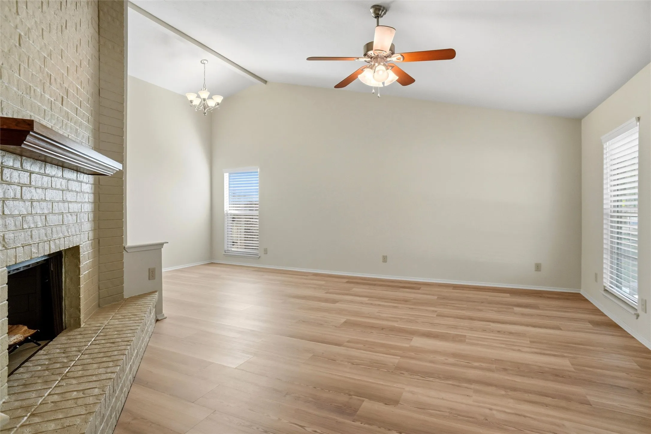 Vaulted ceiling with a contemporary chandelier, wood-finish flooring, brick fireplace with mantel, and window with blinds