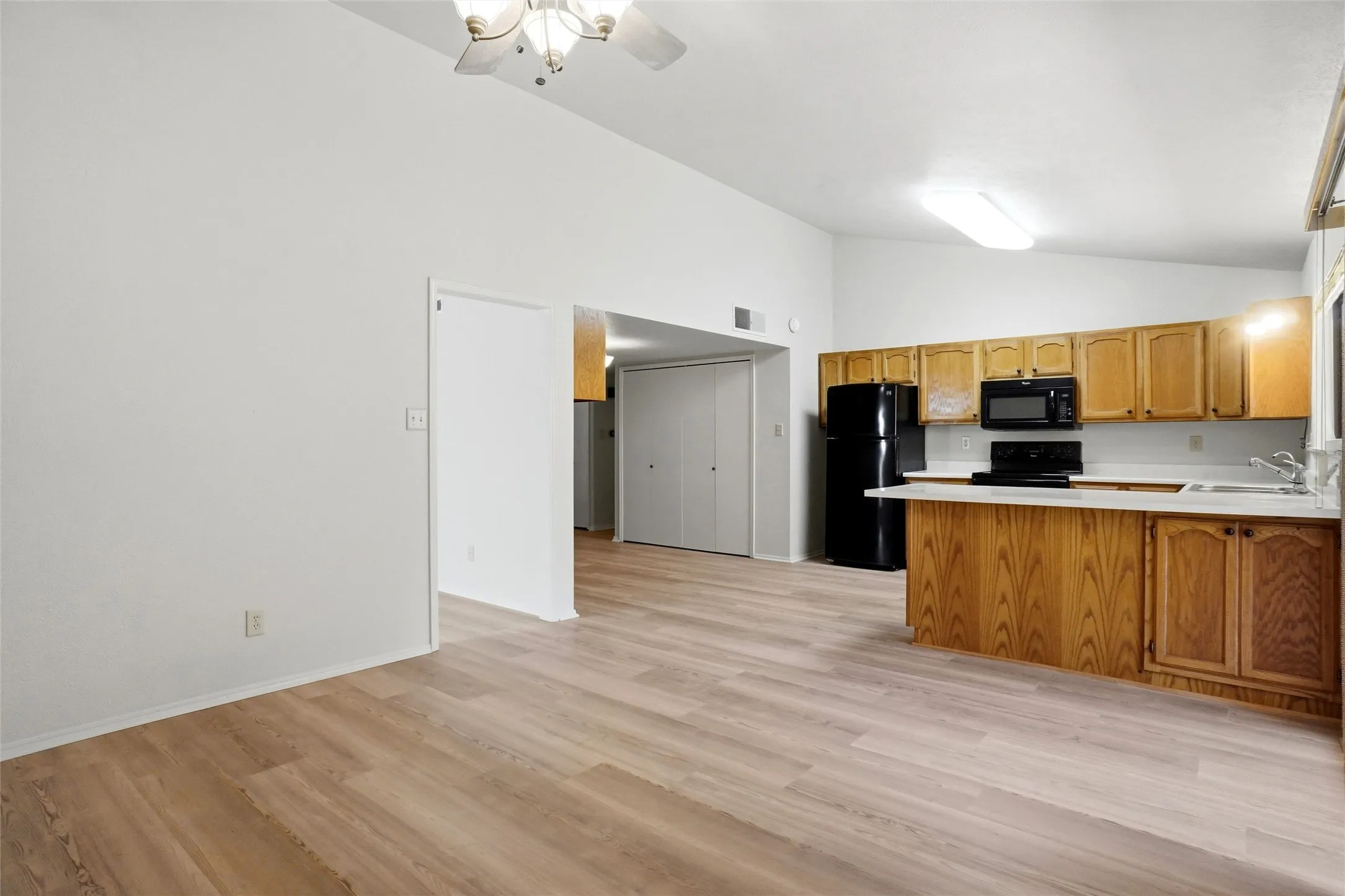Vaulted ceiling with ceiling fan, wood-finish flooring, kitchen with light wood cabinetry, white countertops, and black appliances