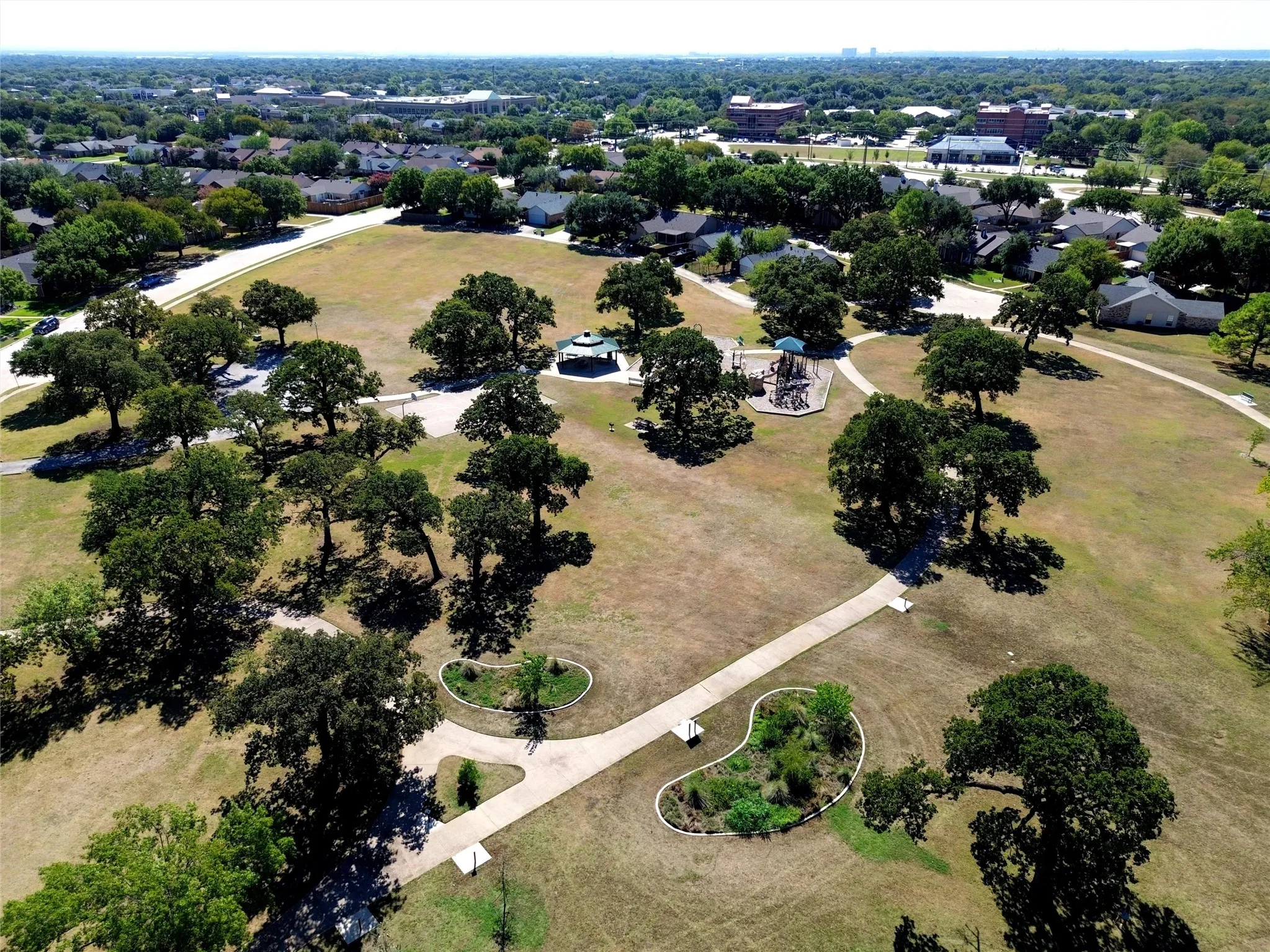 Expansive parkland with mature trees, paved walking paths, a playground structure, and a covered gazebo