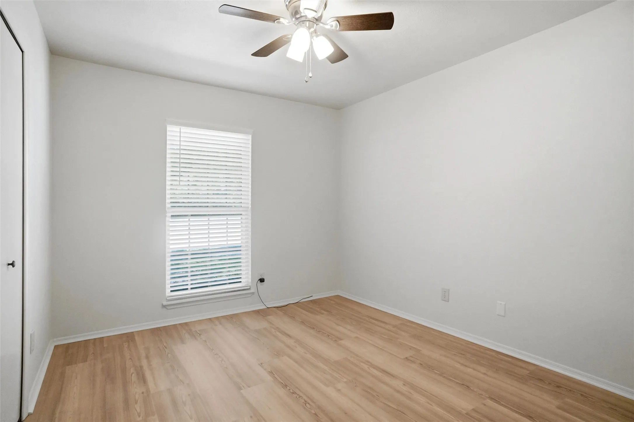 Hardwood-finish flooring, single window with blinds, ceiling fan with light fixture, white walls, and a white door with a black handle
