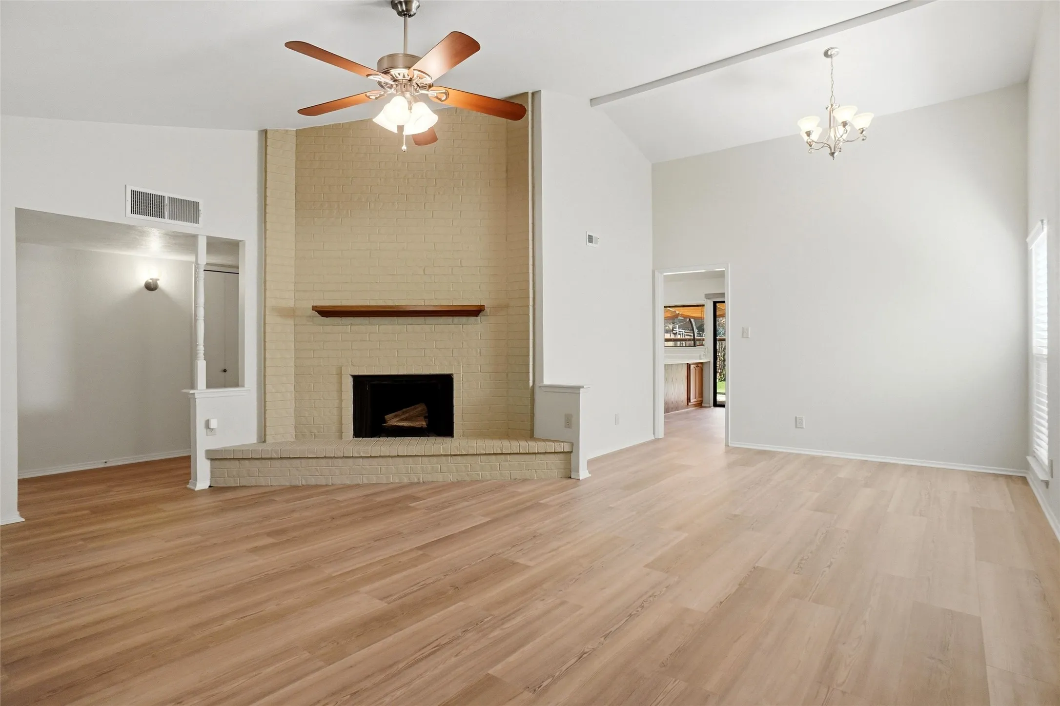 Vaulted ceiling with exposed beam and chandelier, wood-finish flooring, brick fireplace with wood mantel, ceiling fan with integrated lighting, and window with blinds