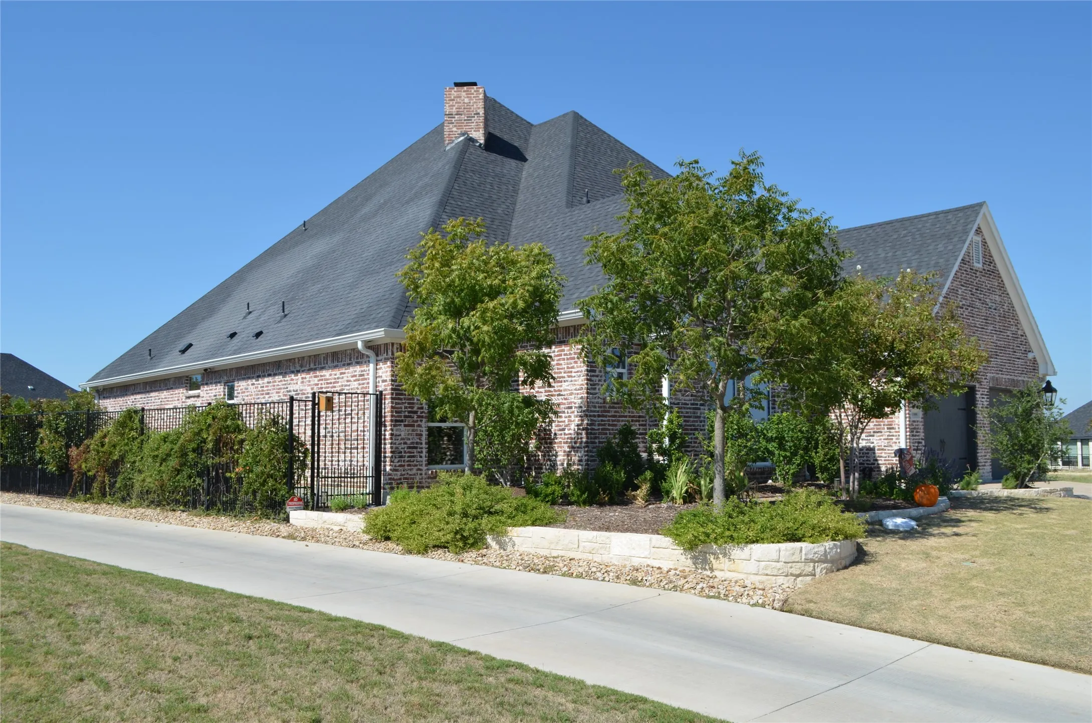 Brick exterior with a dark-shingle roof, featuring a drive, and stone retaining walls, complemented by mature landscaping and a black metal fence