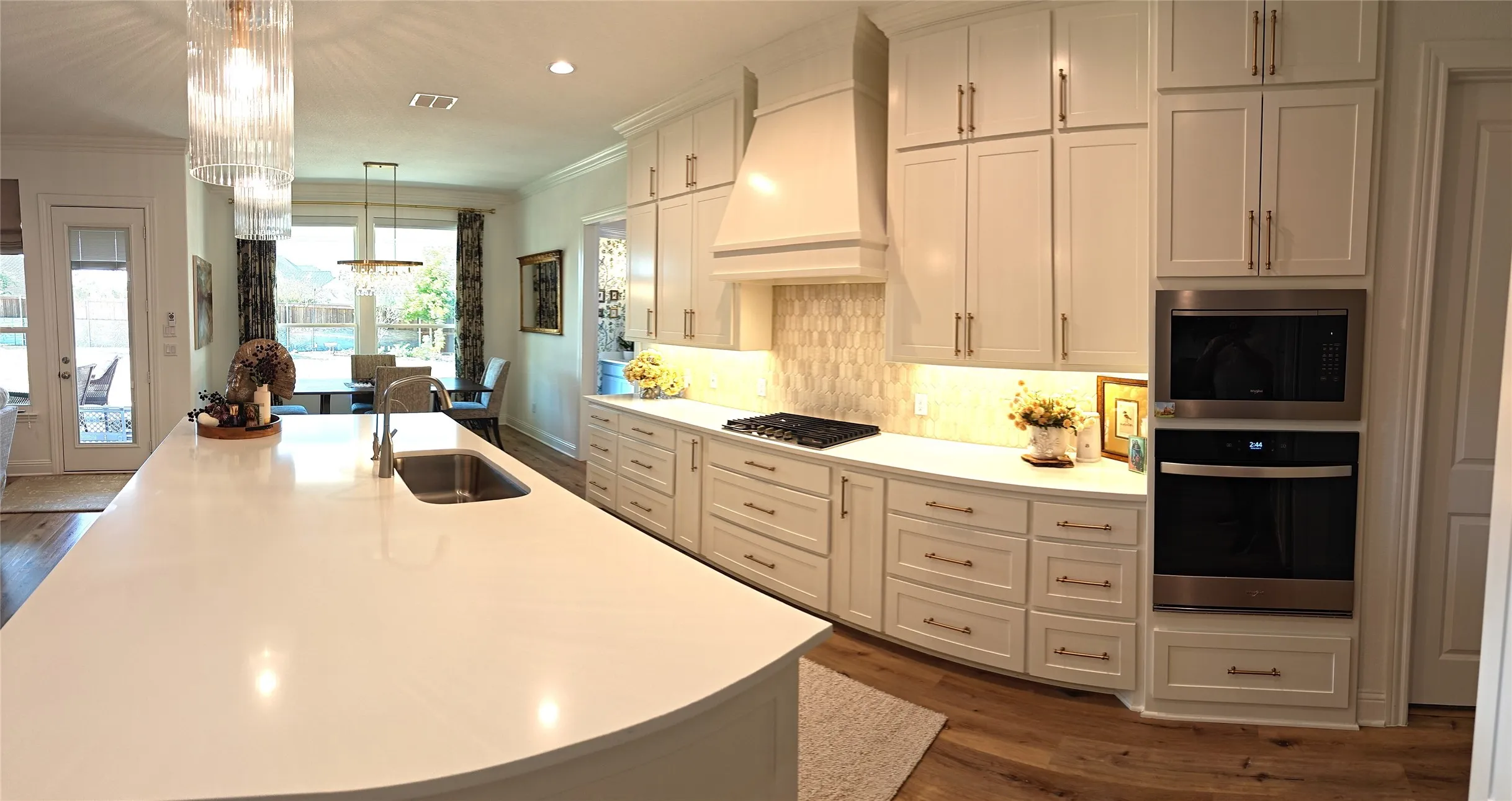 Spacious kitchen featuring an expansive island with an undermount sink, white cabinetry with gold-toned hardware, built-in stainless steel appliances, and wood-finish flooring