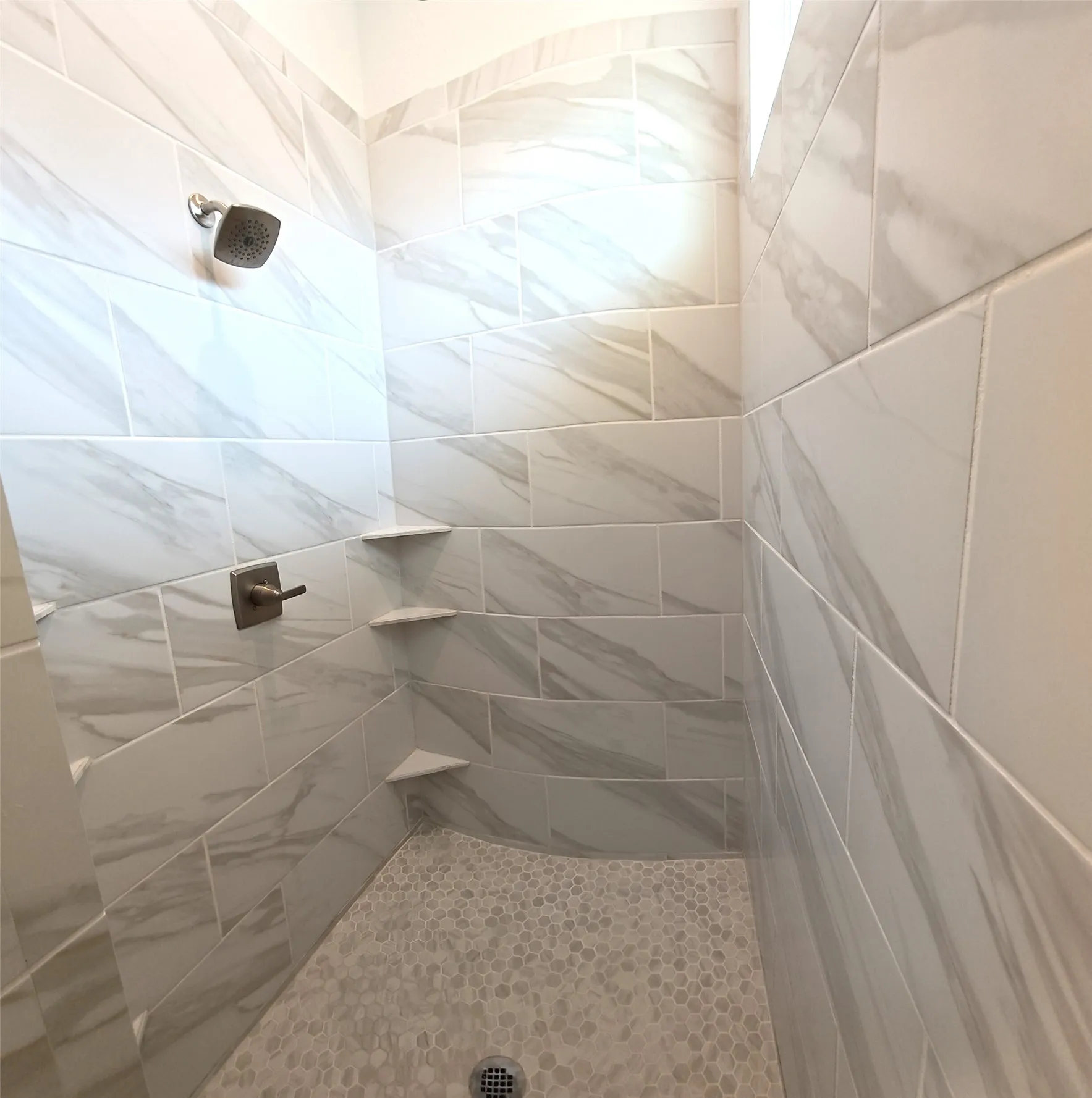 Shower featuring large format wall tiles, pebble-style floor tiling, integrated corner shelving, and a showerhead with a bronze-finish handle