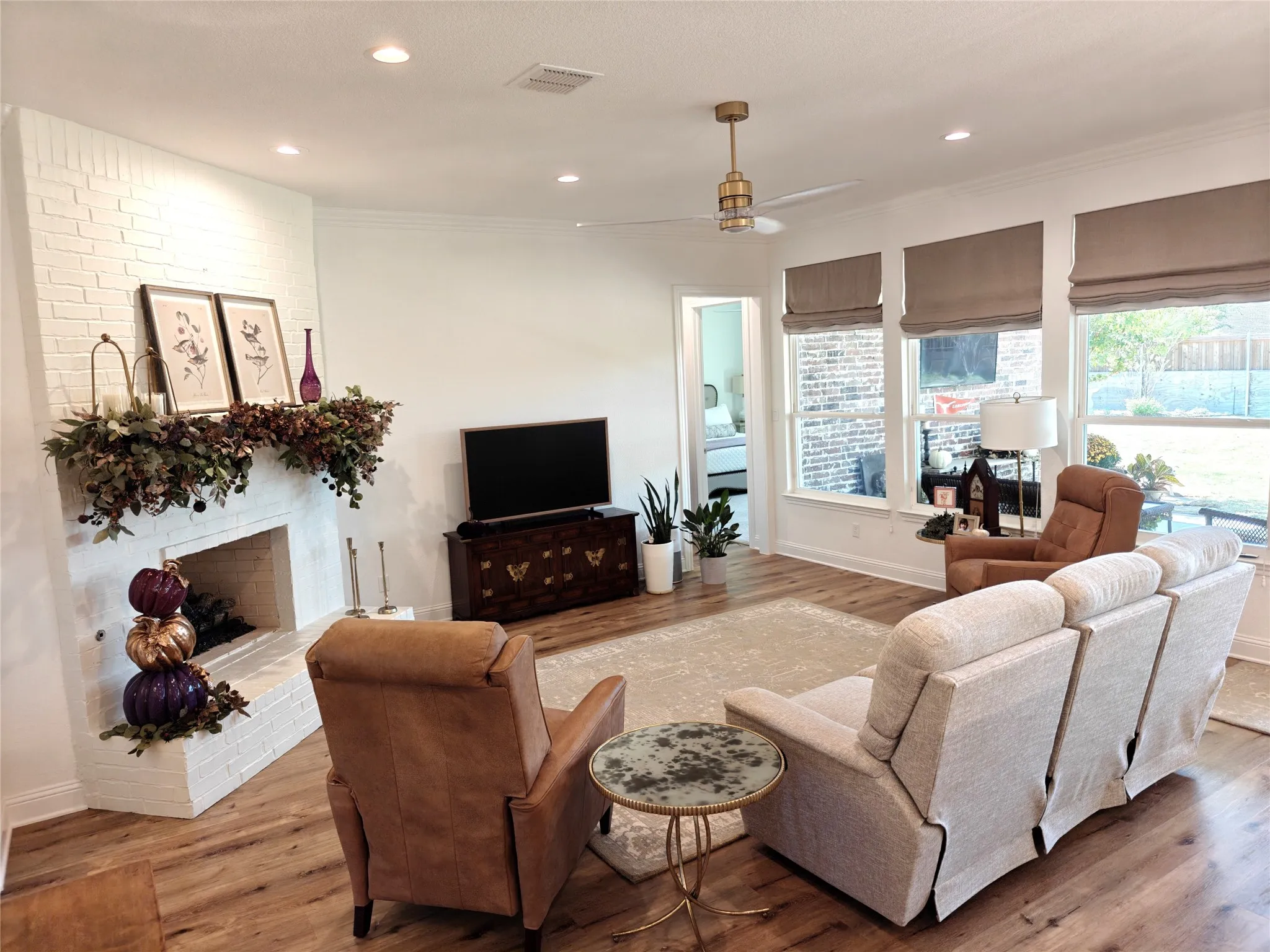 Spacious living area featuring a white brick fireplace, wood-finish flooring, recessed lighting, and multiple large windows with Roman shades