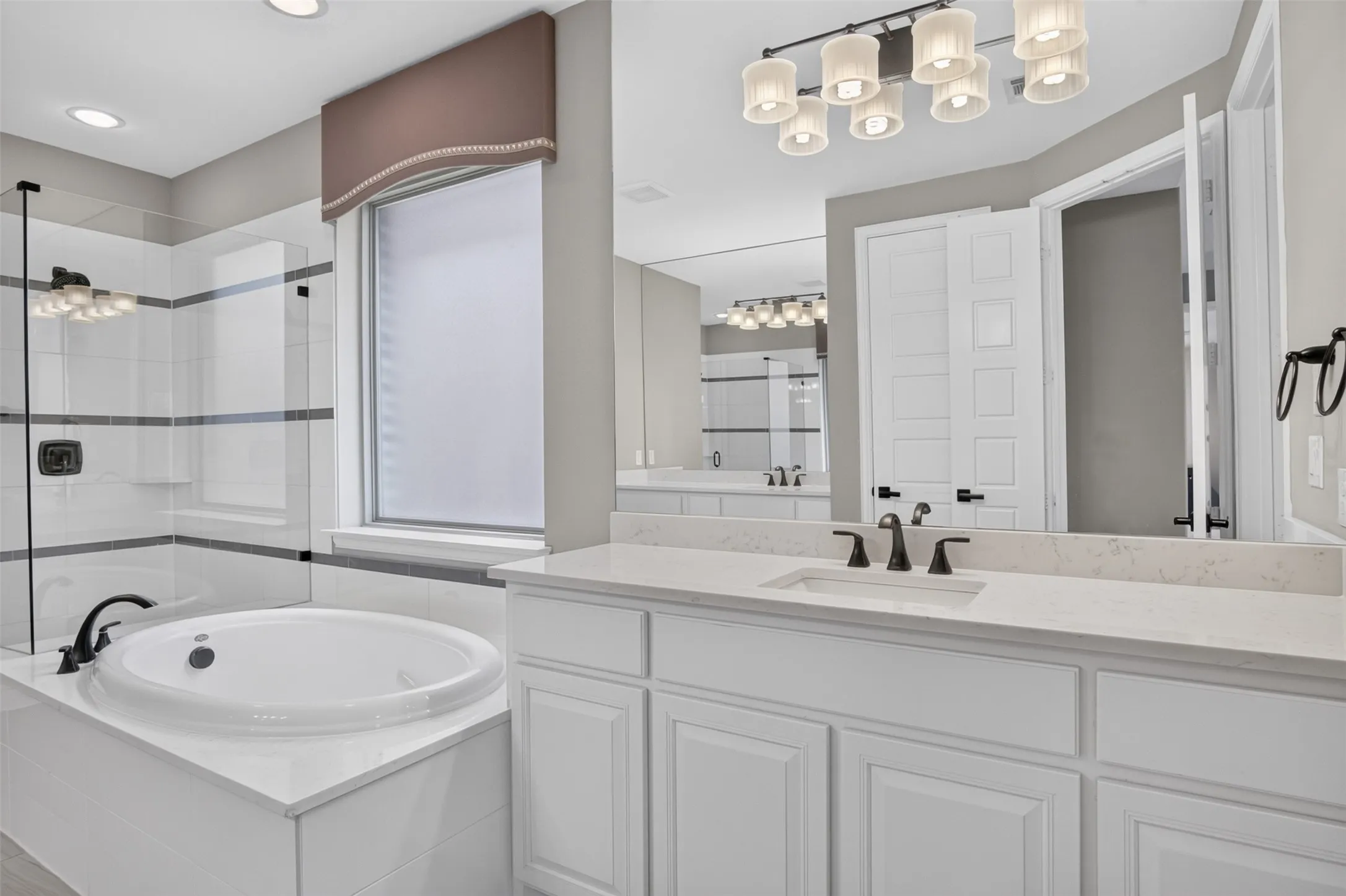 Built-in soaking tub with surround tiling, glass-enclosed shower, white vanity with stone countertop, undermount sink, and dark bronze fixtures