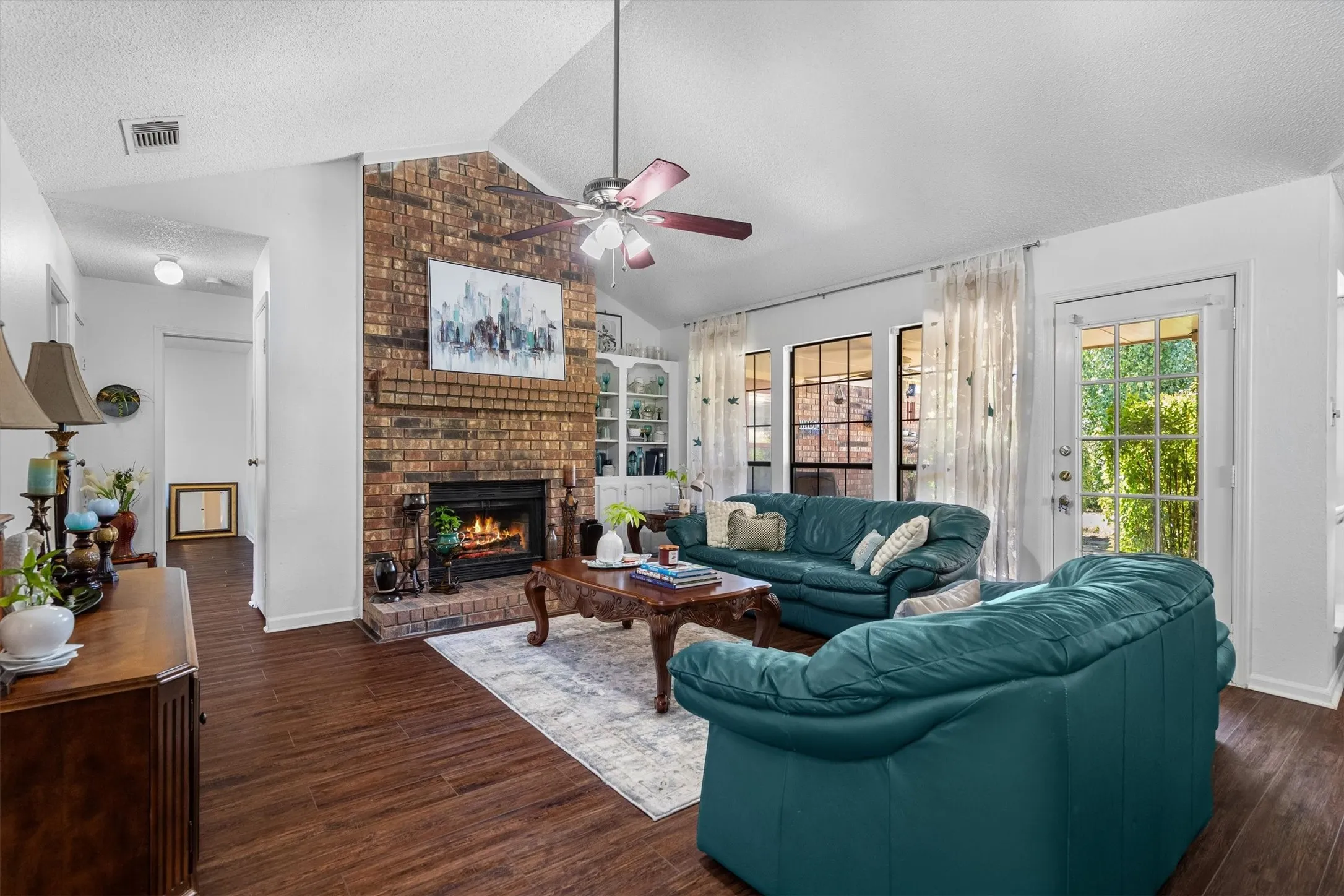 Vaulted ceiling, brick fireplace with mantel, dark wood-finish flooring, ceiling fan, and backyard access via a glass-paneled door