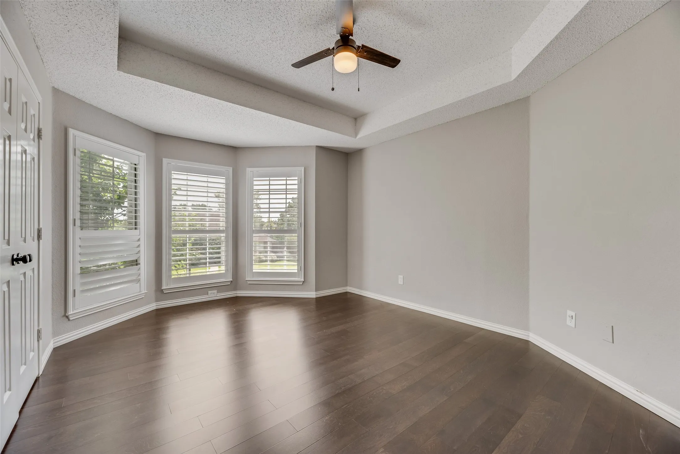Recessed ceiling with a ceiling fan, bay window with plantation shutters, light gray wall paint, and dark wood-finish flooring