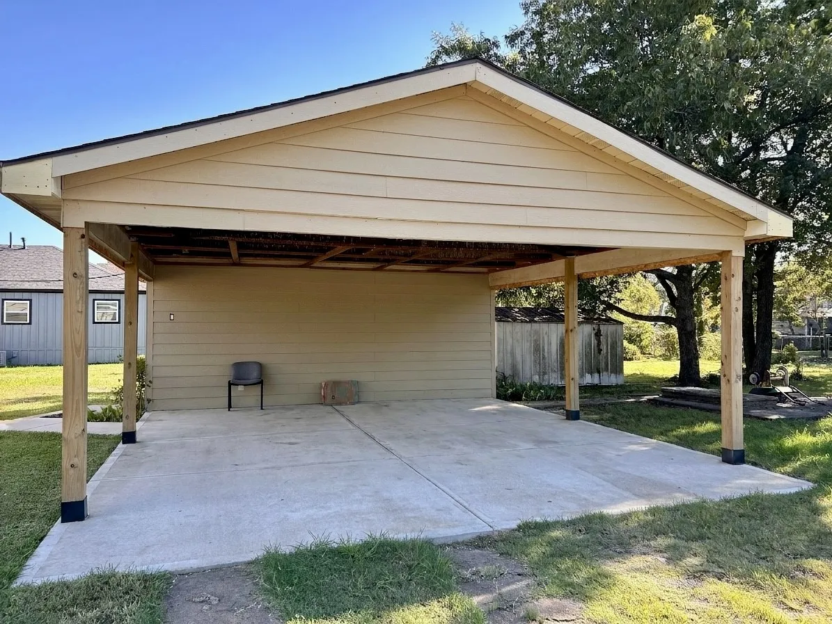 Carport structure with a pitched roof, light-colored siding, exposed wood support beams, and a concrete pad