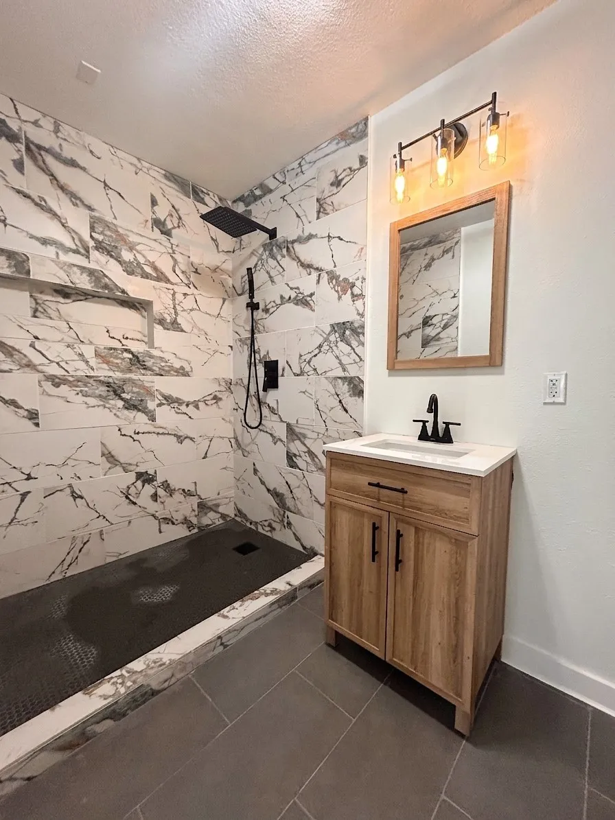 Walk-in shower with large format marble-look tile and matte black fixtures, wood-finish vanity with a white countertop and rectangular sink, and rectangular floor tiles