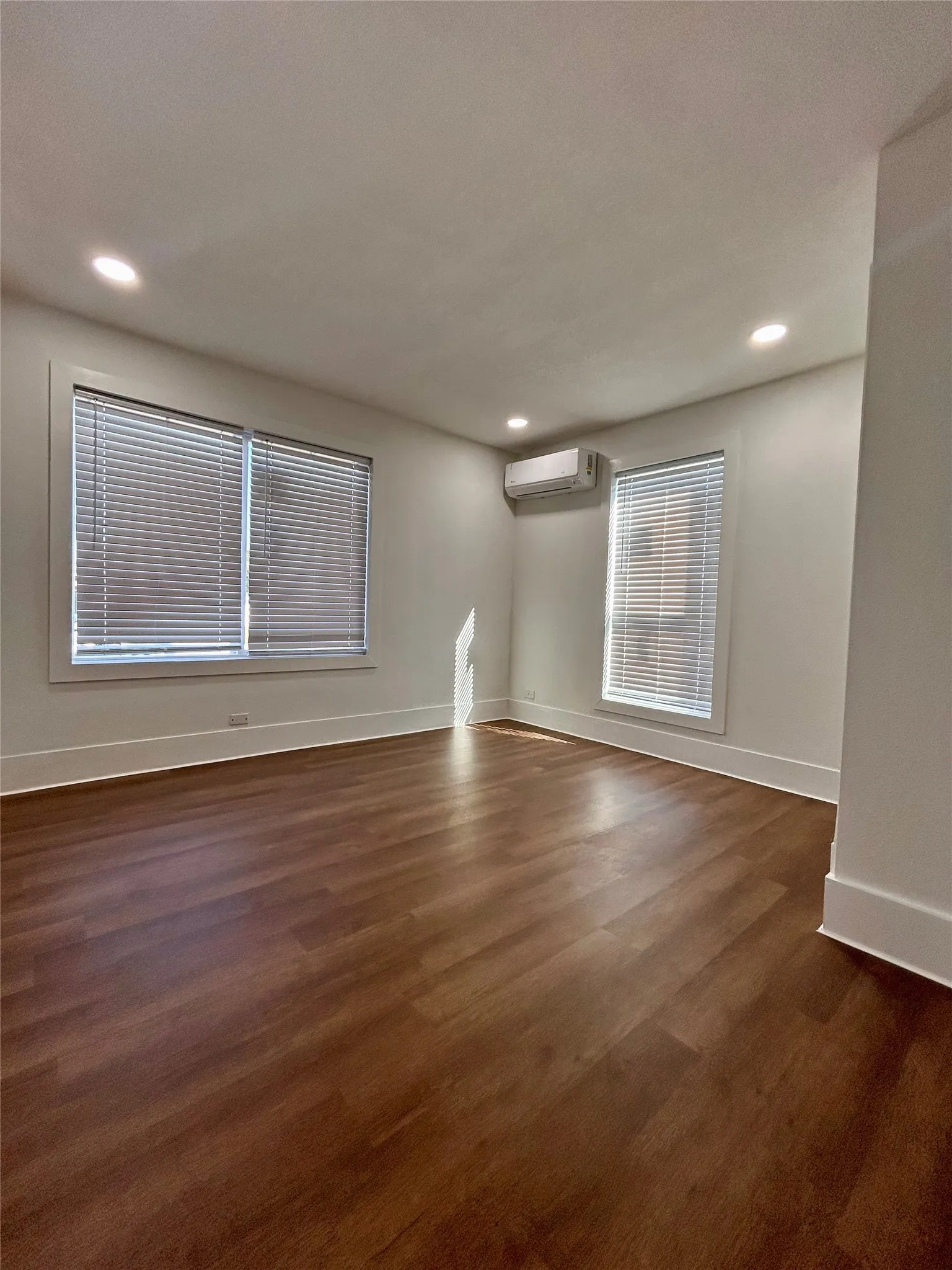 Recessed ceiling lighting, multiple windows with blinds, wall-mounted climate control unit, and wood-finish flooring